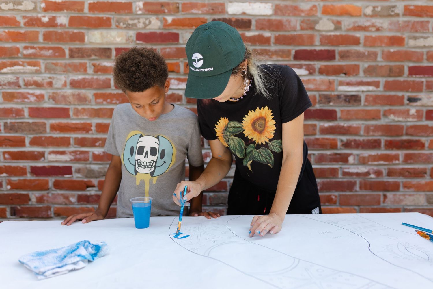 A woman in a green cap and sunflower shirt helps a boy paint on a white canvas outdoors near a brick wall.