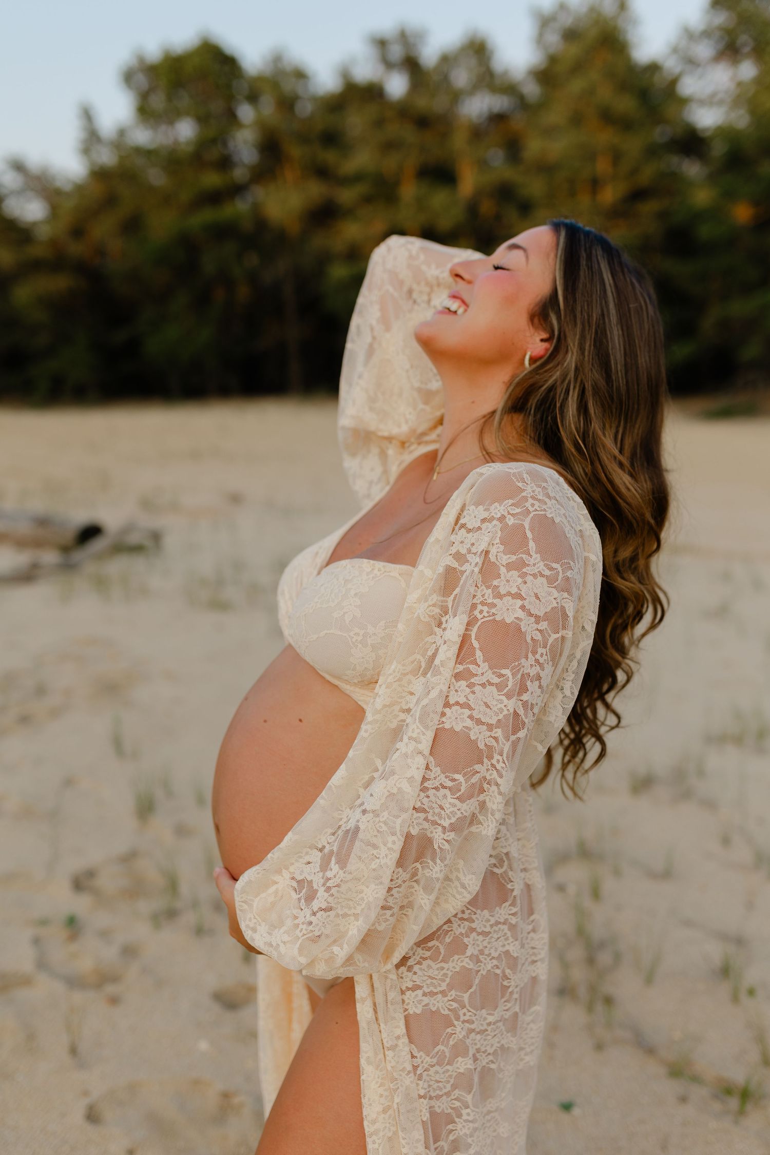 Joyful pregnant woman in lace maternity gown laughs while holding baby bump on sandy beach at sunset.