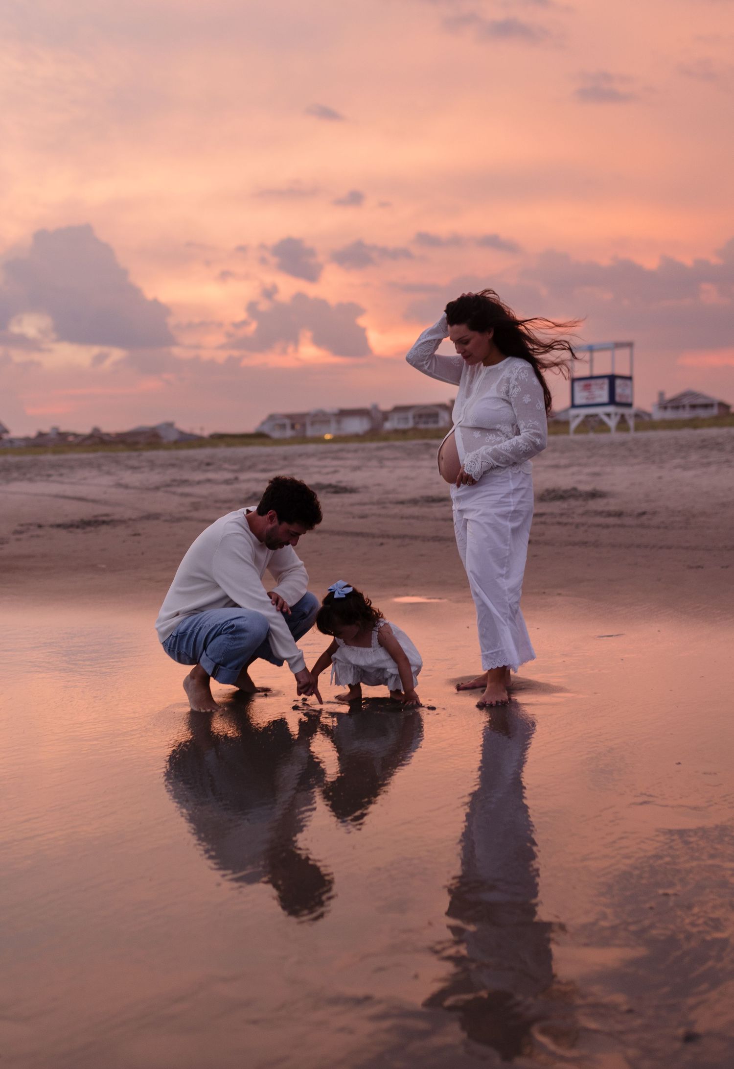 Family maternity photo at sunset on beach, pregnant woman in white watches man and toddler play in wet sand.