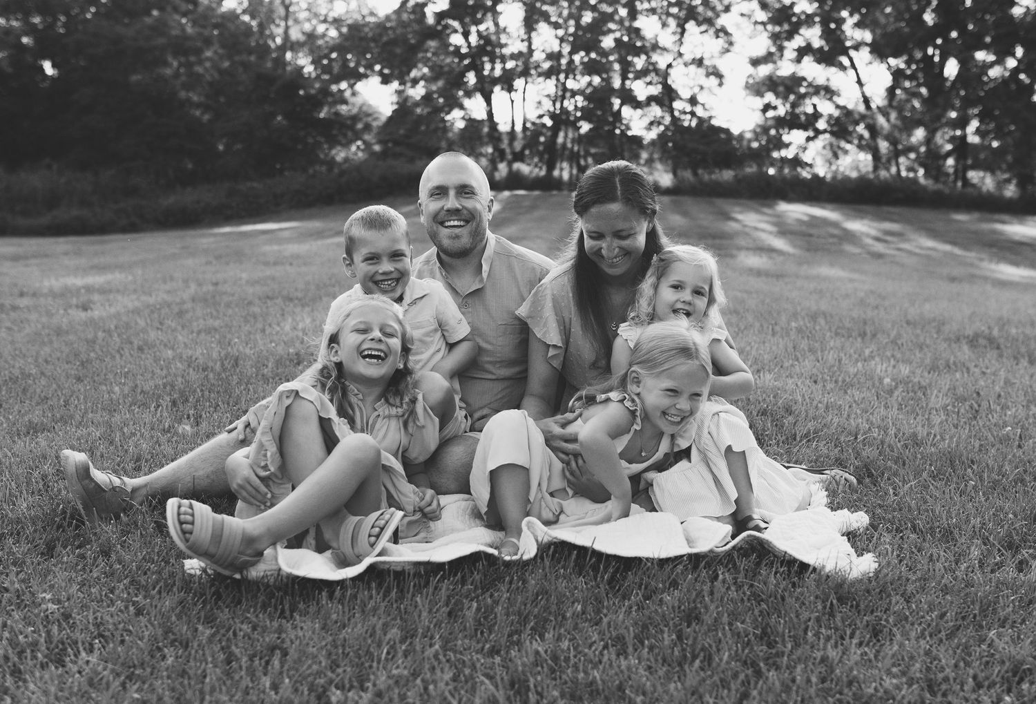 Happy family of six sitting together on a blanket in a grassy field in black and white.