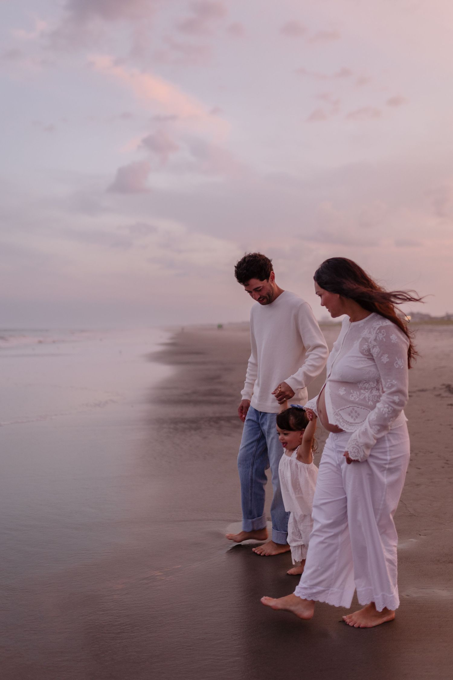 Family of four walking barefoot on a beach at sunset, wearing white outfits, with a pastel pink sky backdrop.