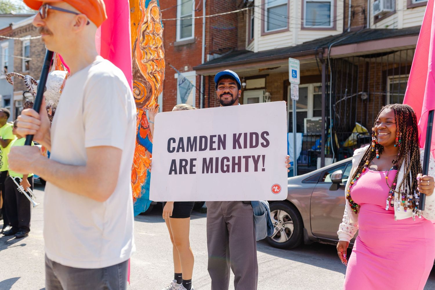 People marching in a Camden parade, holding a sign reading Camden Kids Are Mighty on a sunny day.