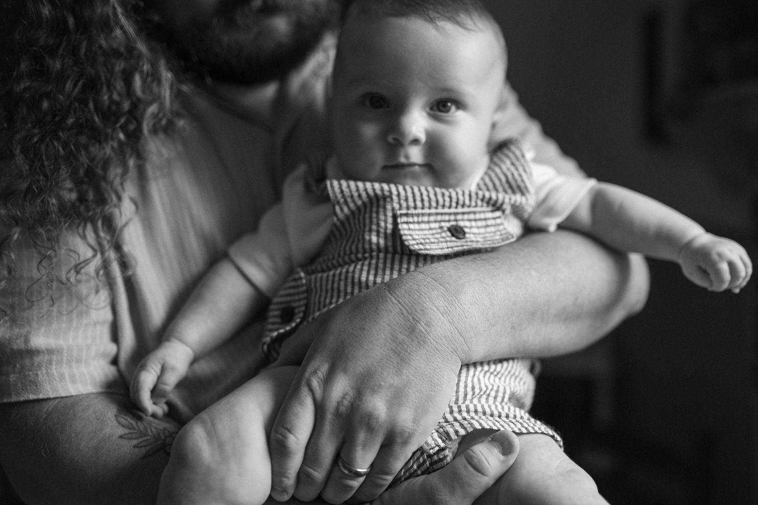 Black and white photo of a bearded father with curly hair cradling his infant in a striped outfit.