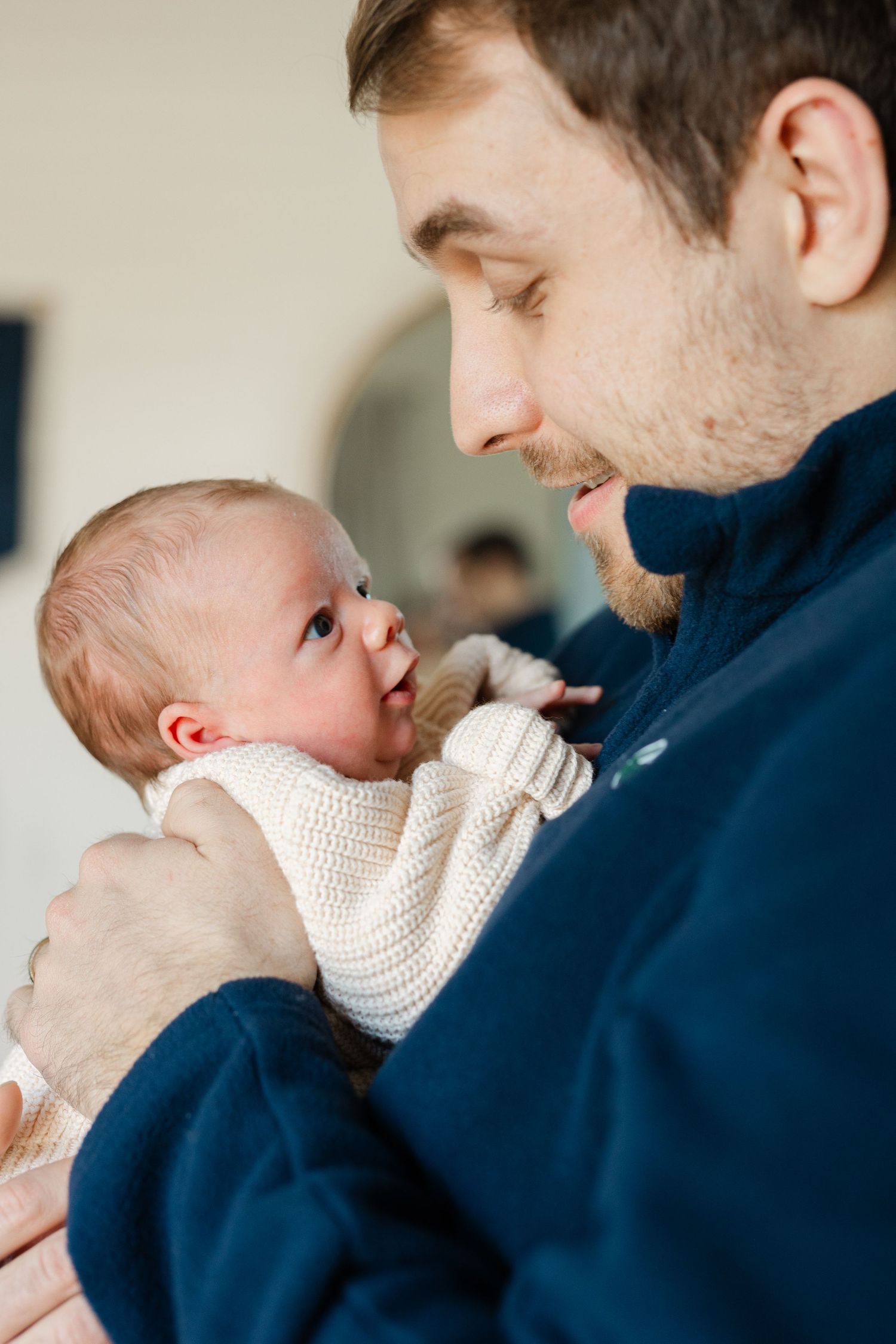Father in navy jacket tenderly holds newborn baby in cream knit sweater, sharing a gentle moment.