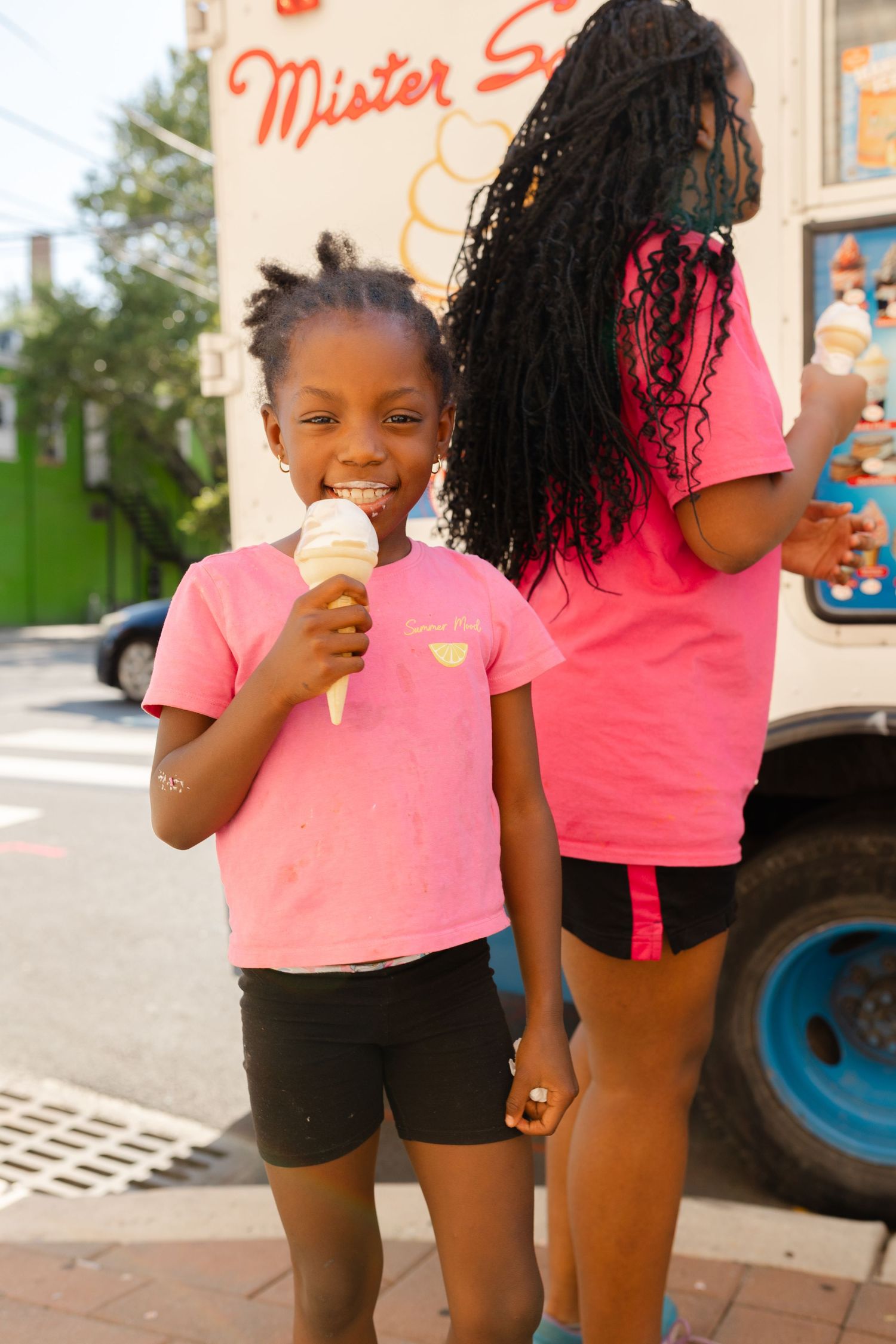 Two young girls in pink shirts enjoying ice cream cones from a Mister Softee truck on a sunny day.