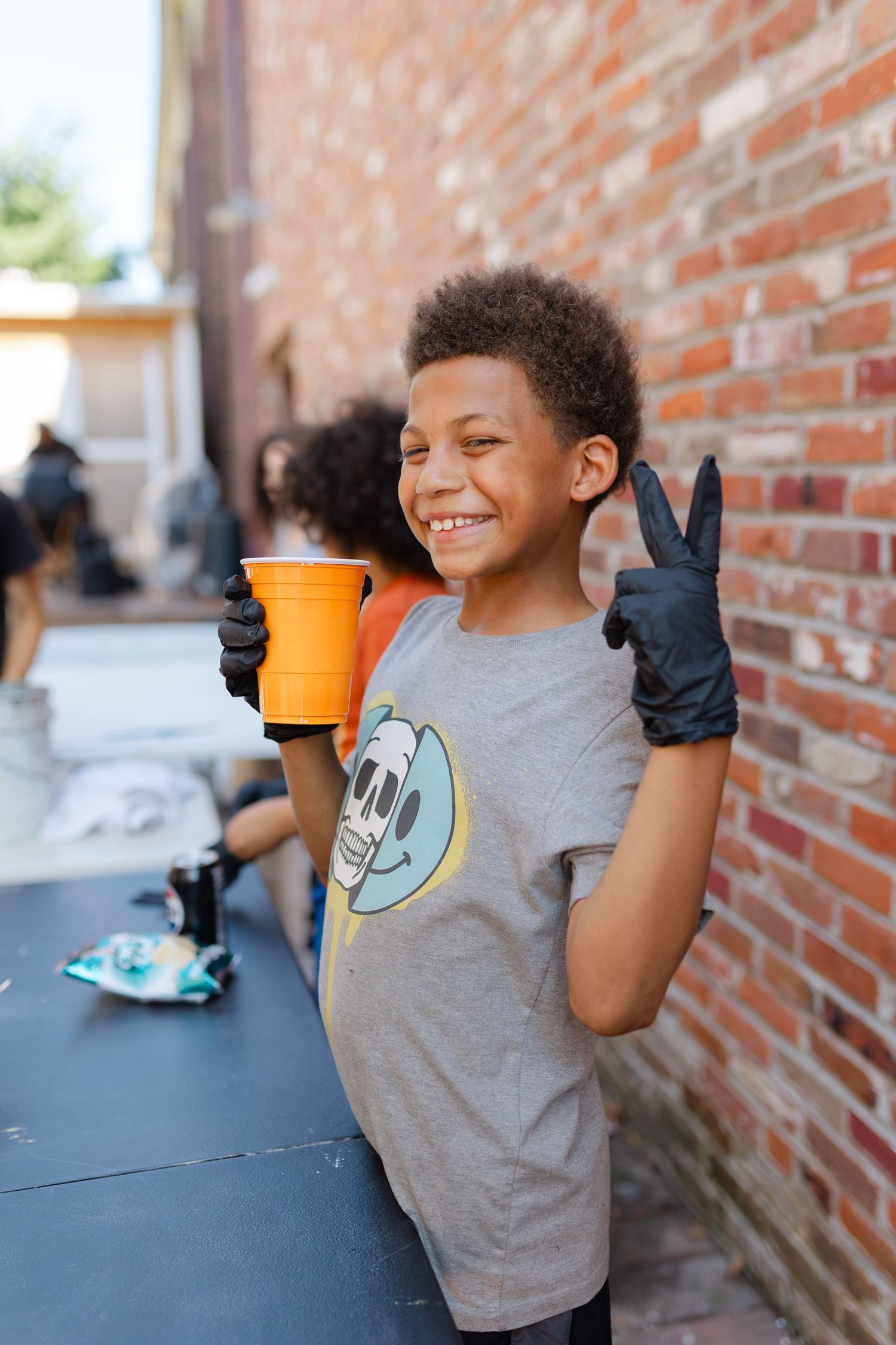 Smiling boy in gray t-shirt wearing black gloves, holding orange cup and making peace sign near brick wall.
