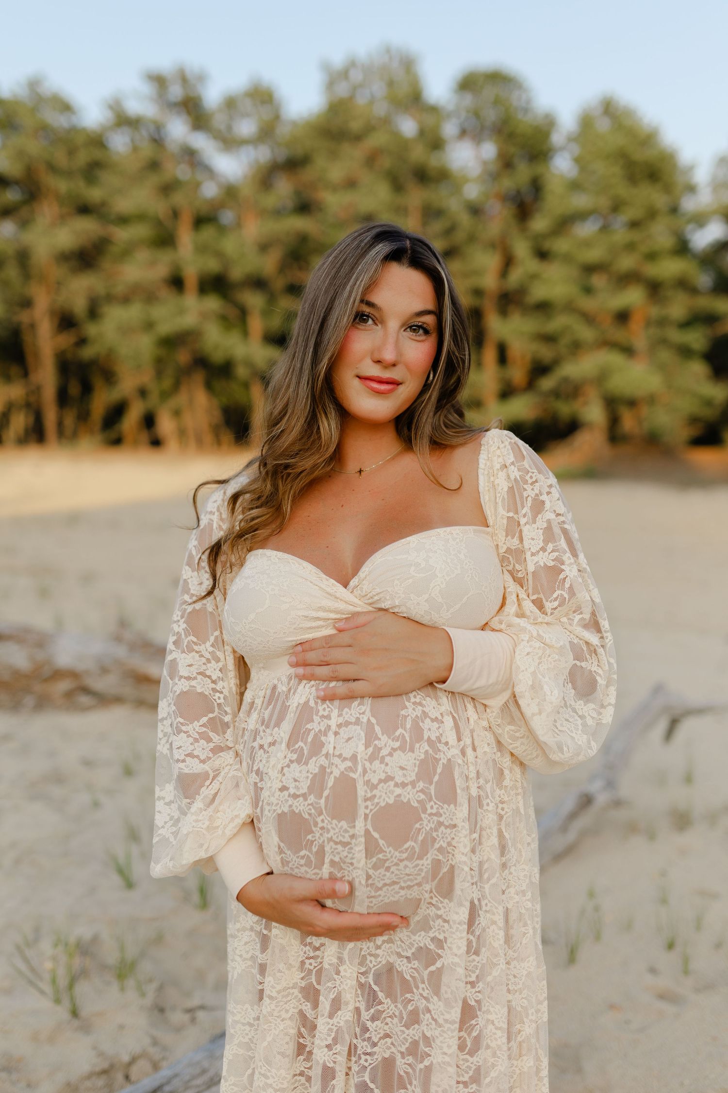 Smiling pregnant woman in cream lace maternity gown holds baby bump on sandy beach at golden hour.