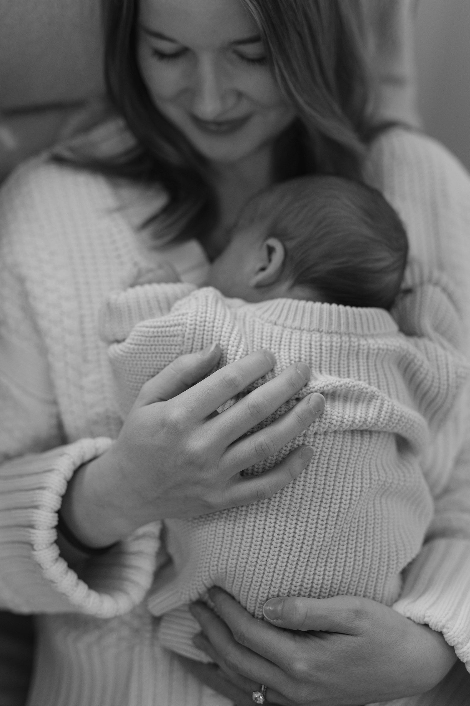 Black and white photo of mother in white knit sweater cradling sleeping newborn baby close to her chest.