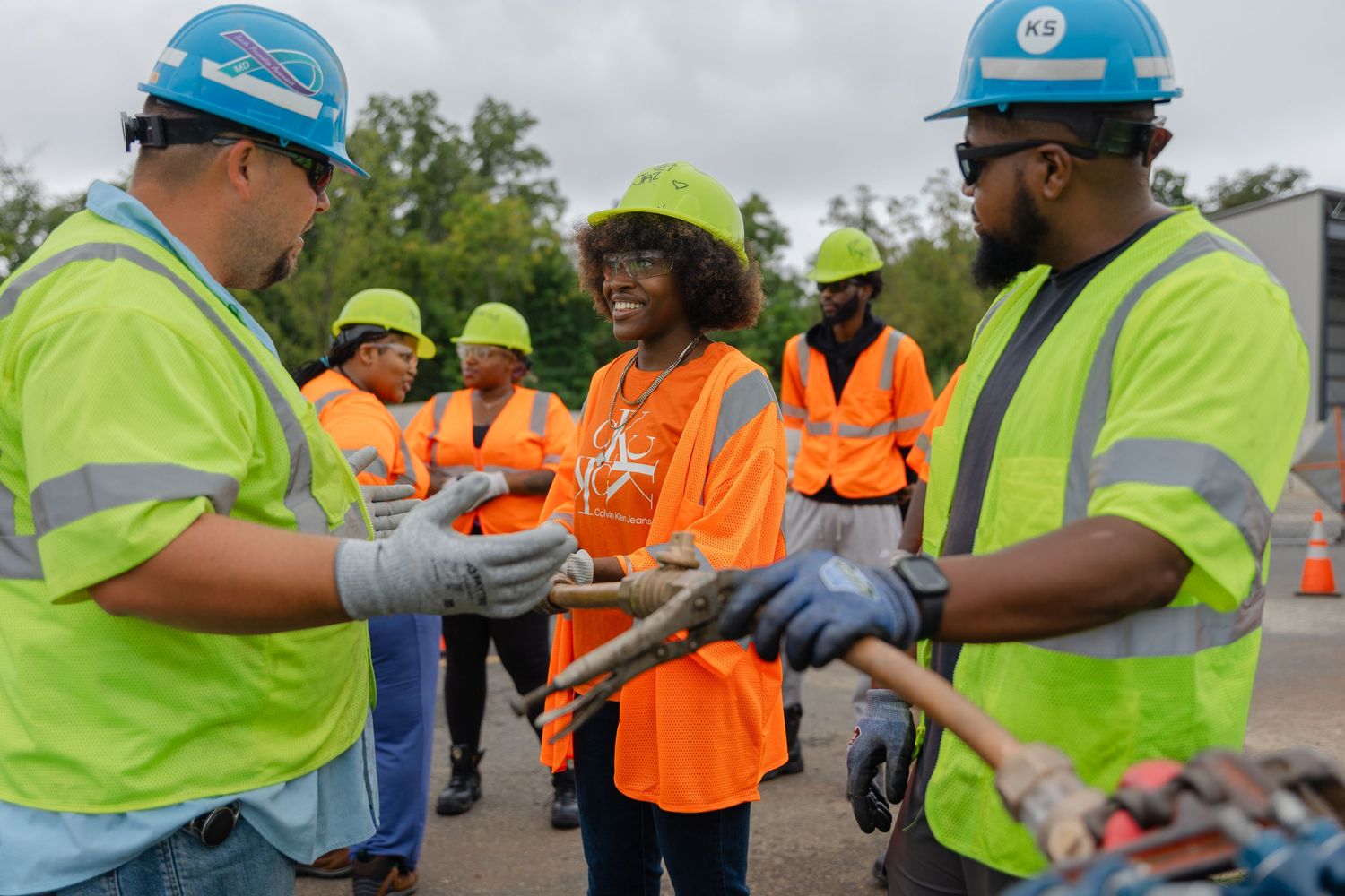 Workers in safety vests and hard hats collaborate on utility cable work outdoors during a team training exercise.