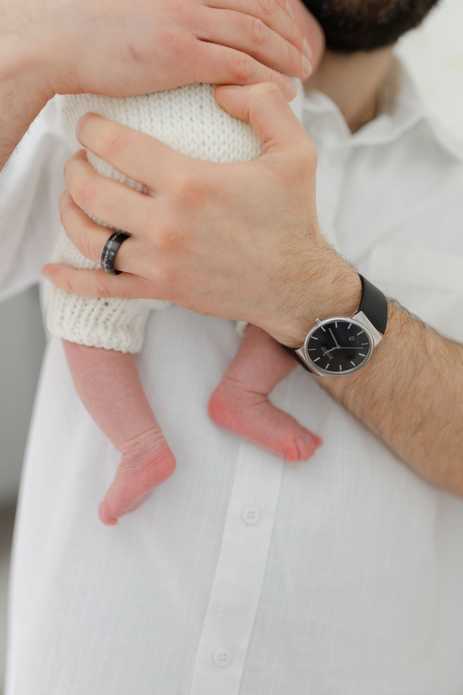 Father holding newborn baby against his chest, wearing a black wedding ring and minimalist watch.