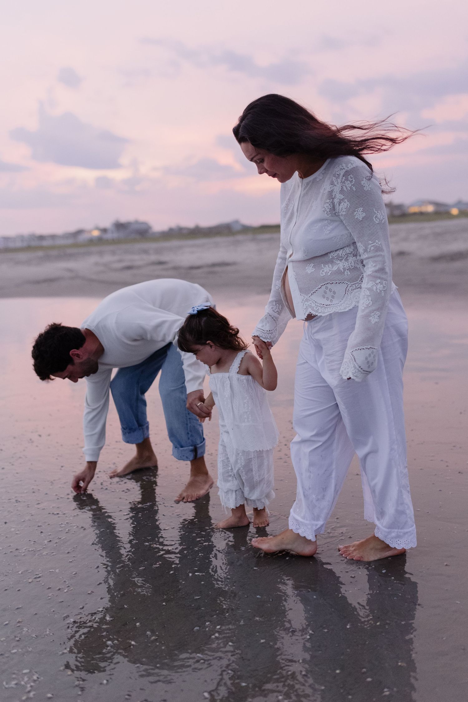 Family of three enjoying a peaceful sunset beach walk, parents guiding toddler along wet sand in matching white outfits.