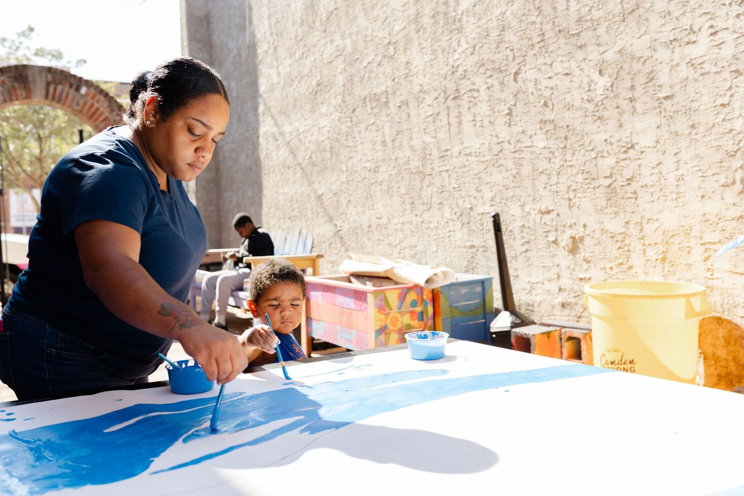 Woman painting blue artwork on table outdoors while a child watches in a sunny courtyard setting.