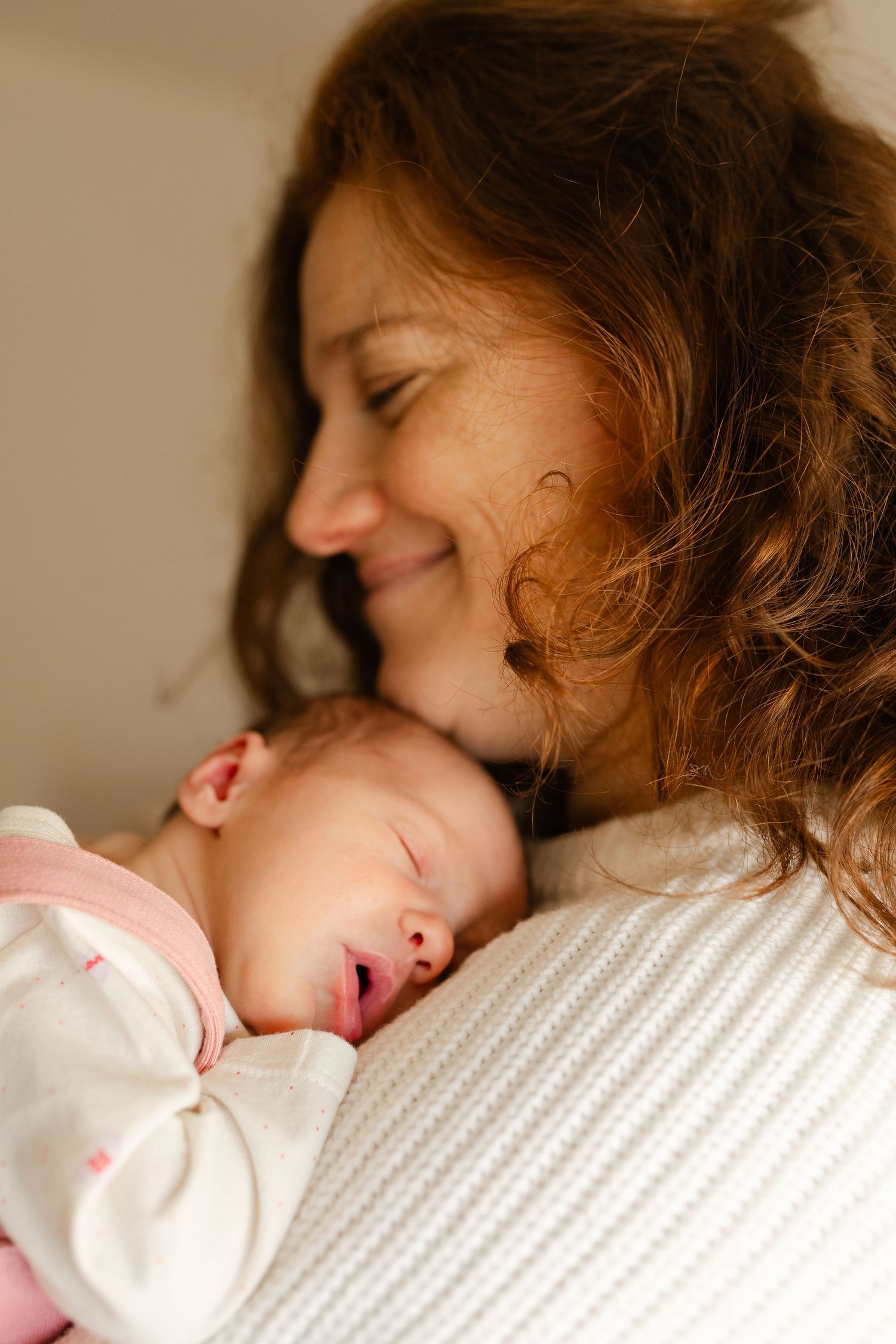 A smiling mother with curly hair gently holds her sleeping newborn baby close to her cheek.