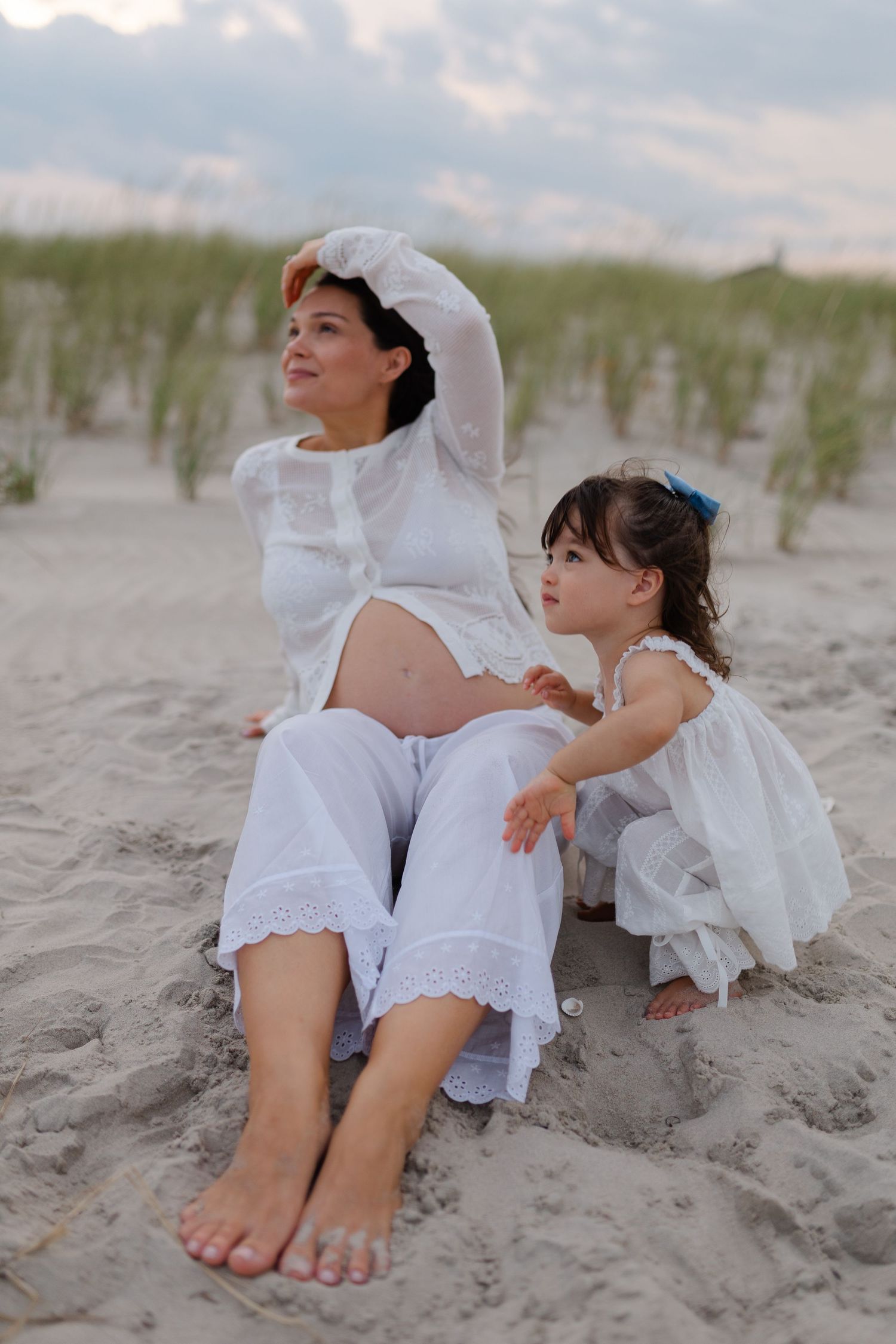 Pregnant woman in white sits on beach sand with toddler girl in white dress, both gazing into the distance.
