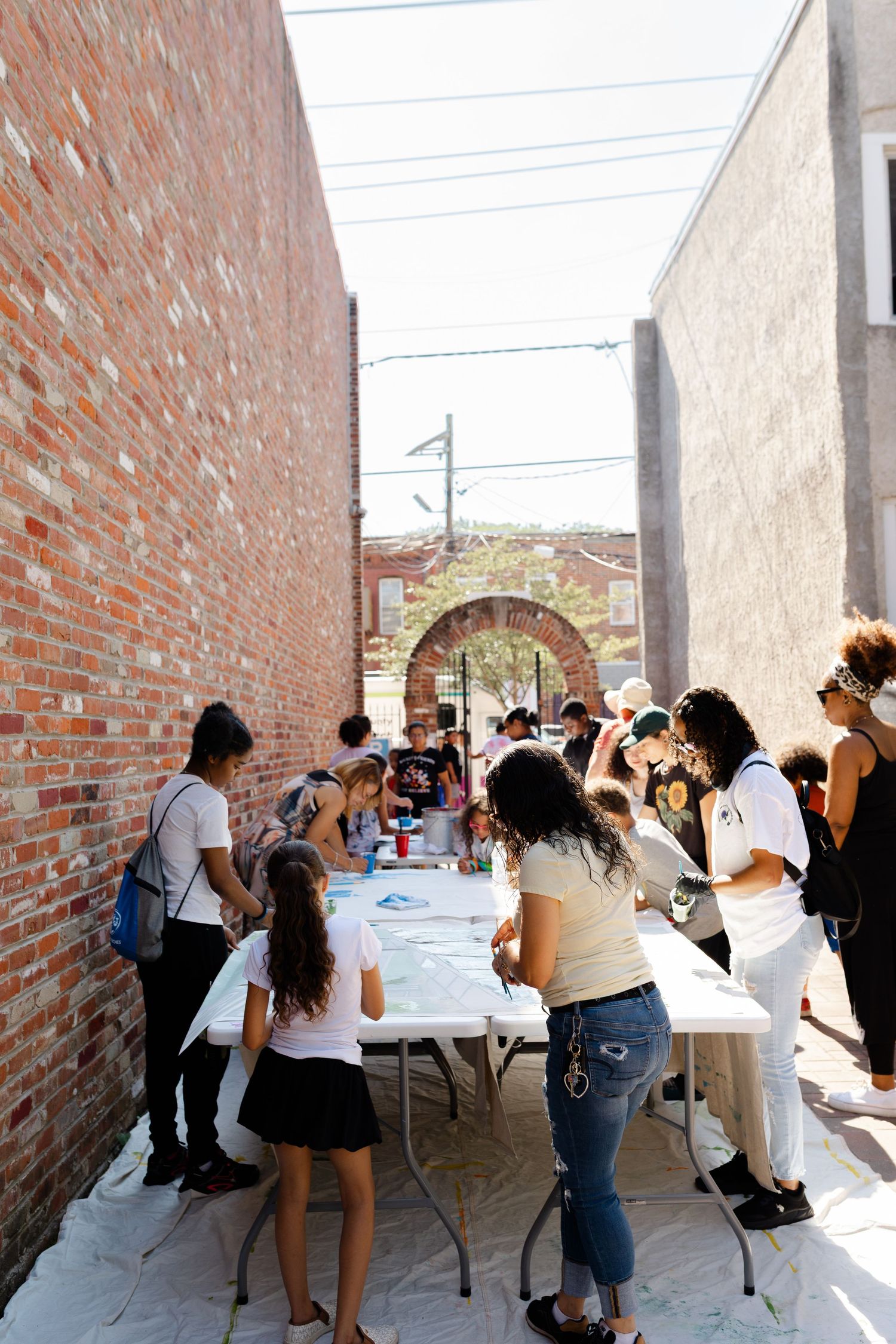 People gather around white tables in a sunlit urban alley between brick buildings for an outdoor community art event.