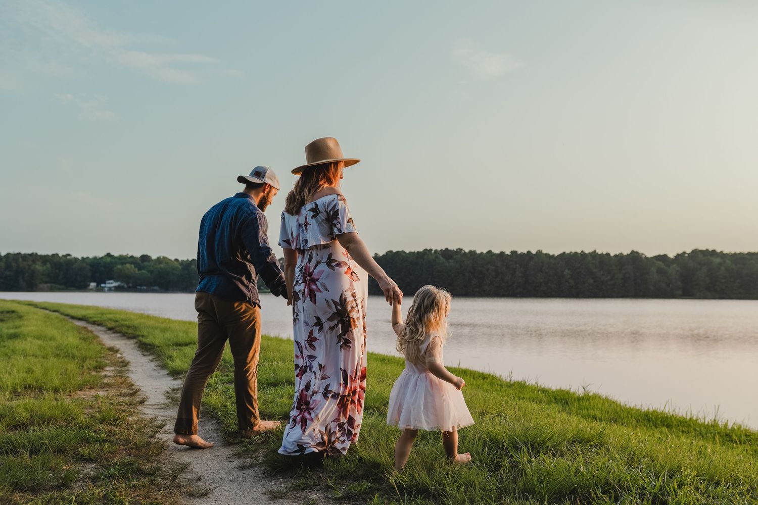 Family of three holding hands by a serene lake at sunset, parents in hats with young daughter in white dress.