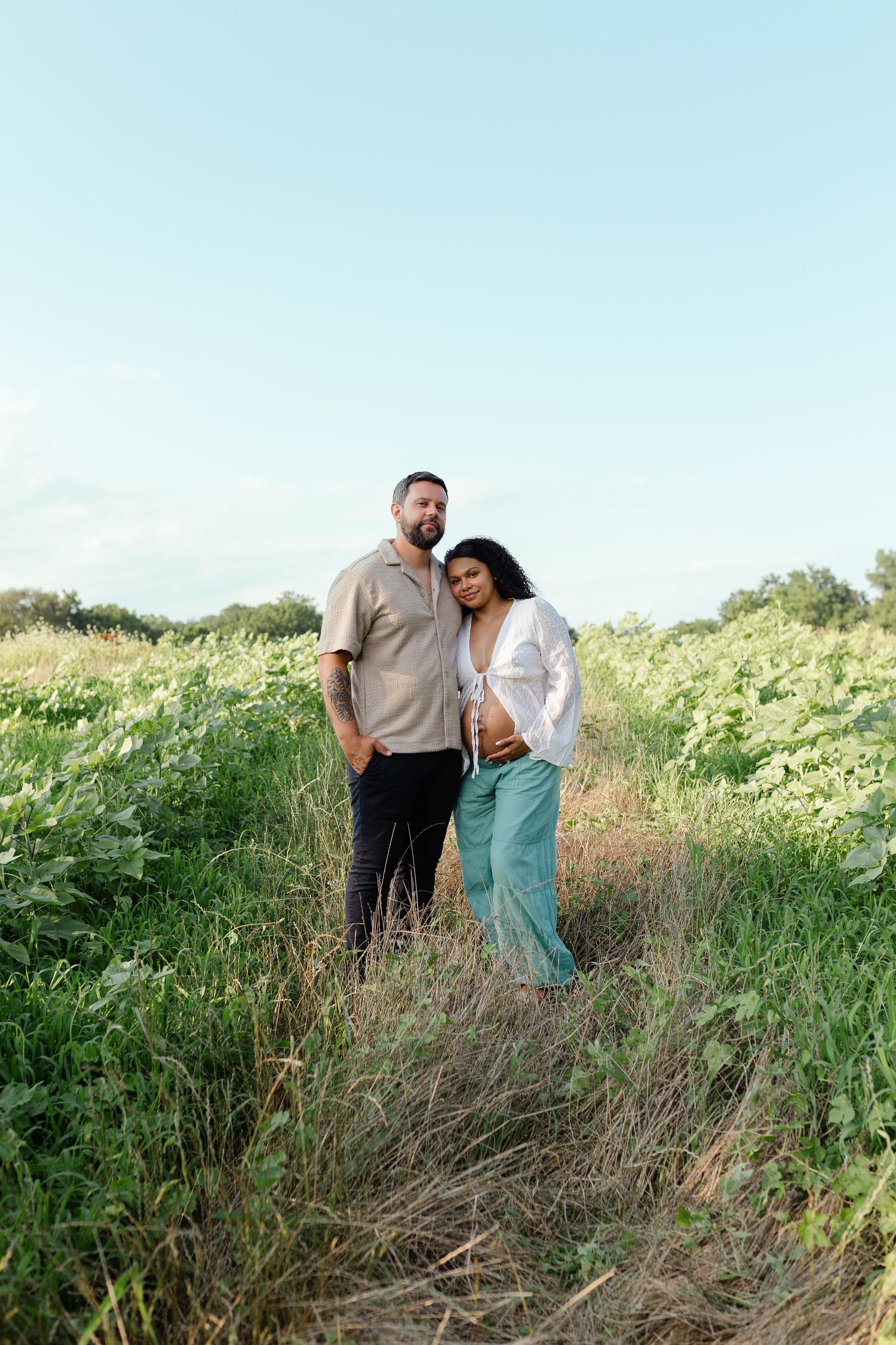 A couple stands together on a grassy field path during an outdoor maternity photo session under a clear sky.
