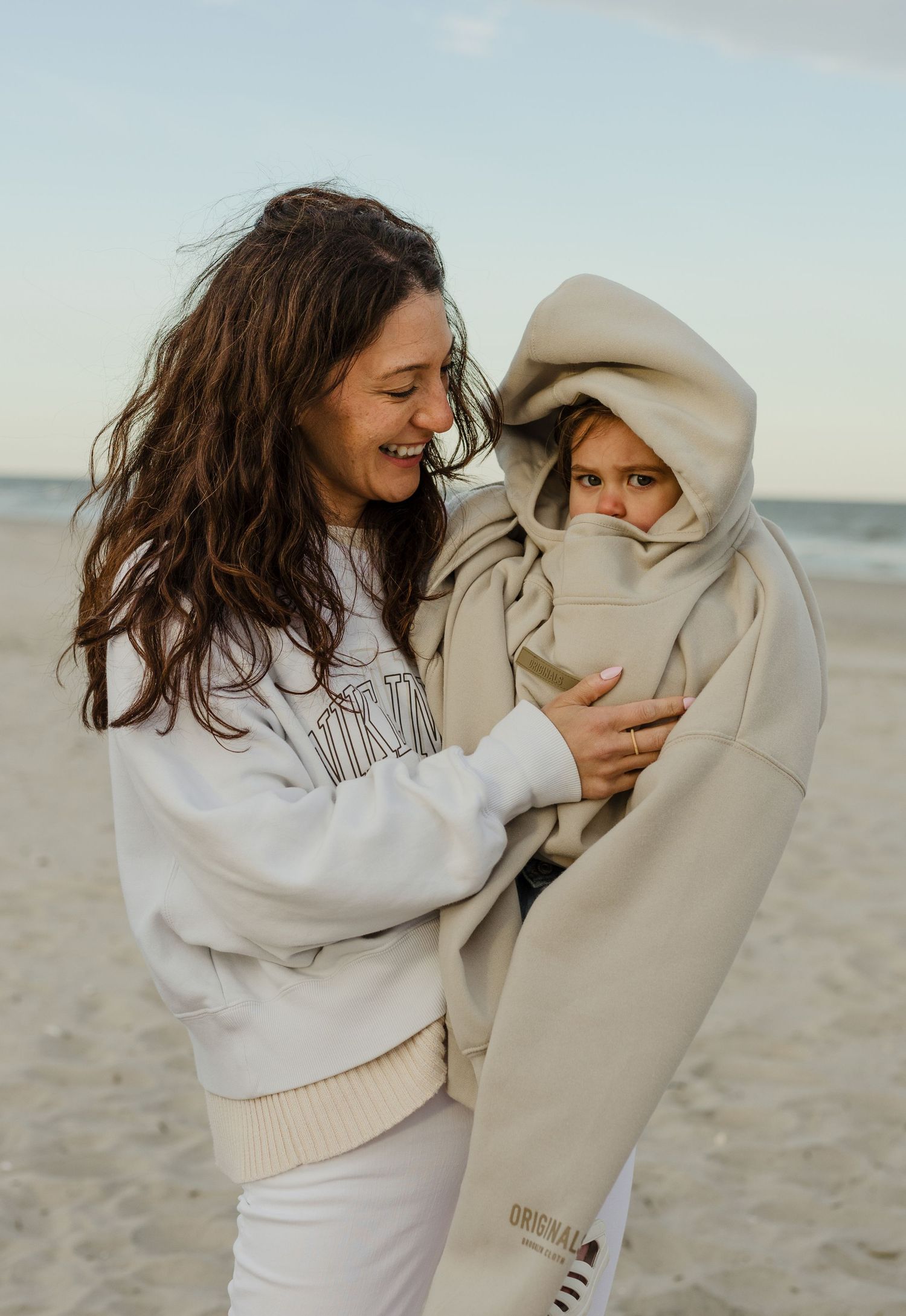 Laughing mother holds child bundled in oversized beige hoodie on a breezy beach.