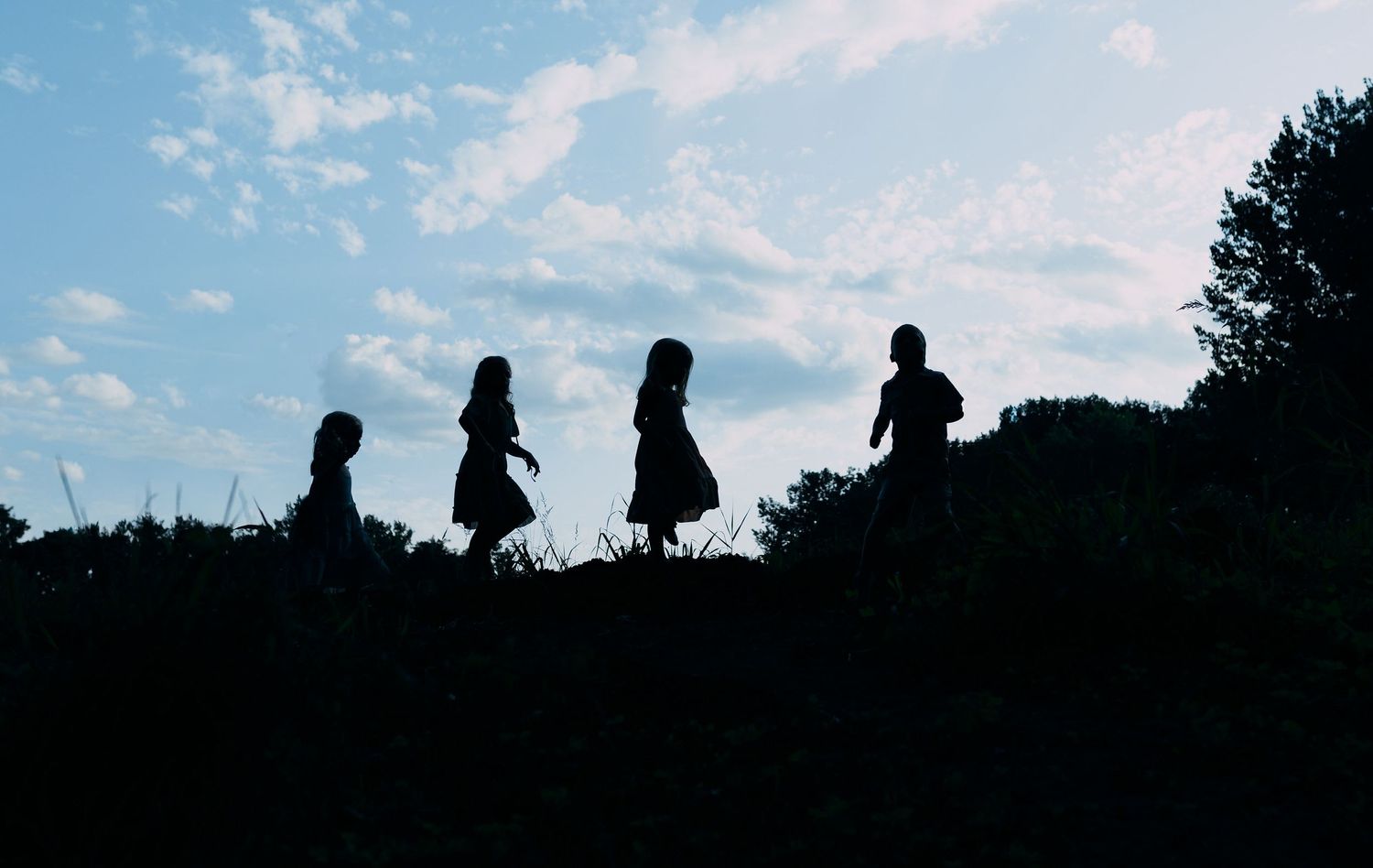 Silhouette of four children walking along a hilltop against a cloudy blue sky.