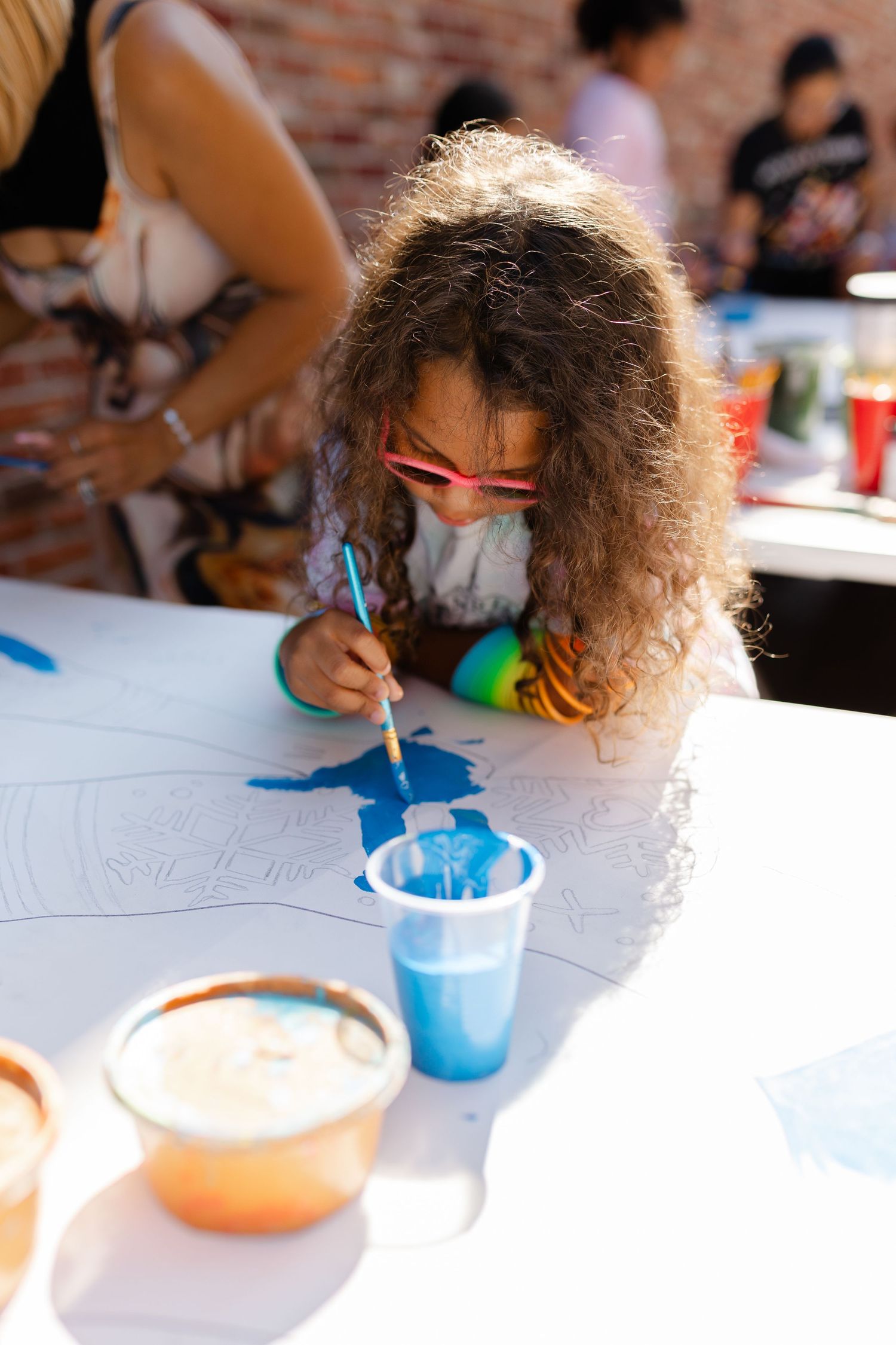 Young girl with curly hair and pink glasses painting blue watercolors on a large white canvas outdoors.