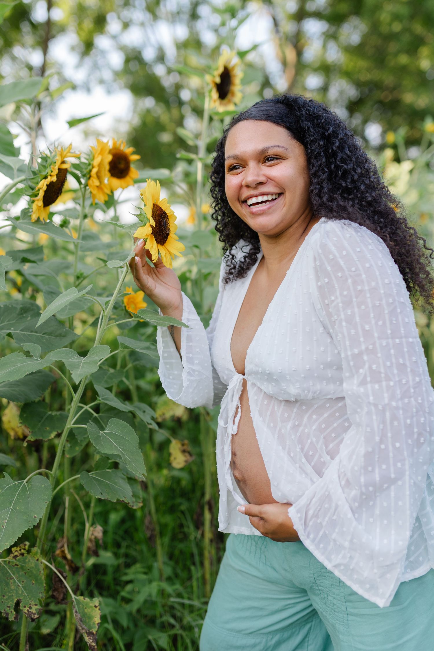 Smiling pregnant woman in white top touching sunflowers in a sunflower field during a maternity photo session.