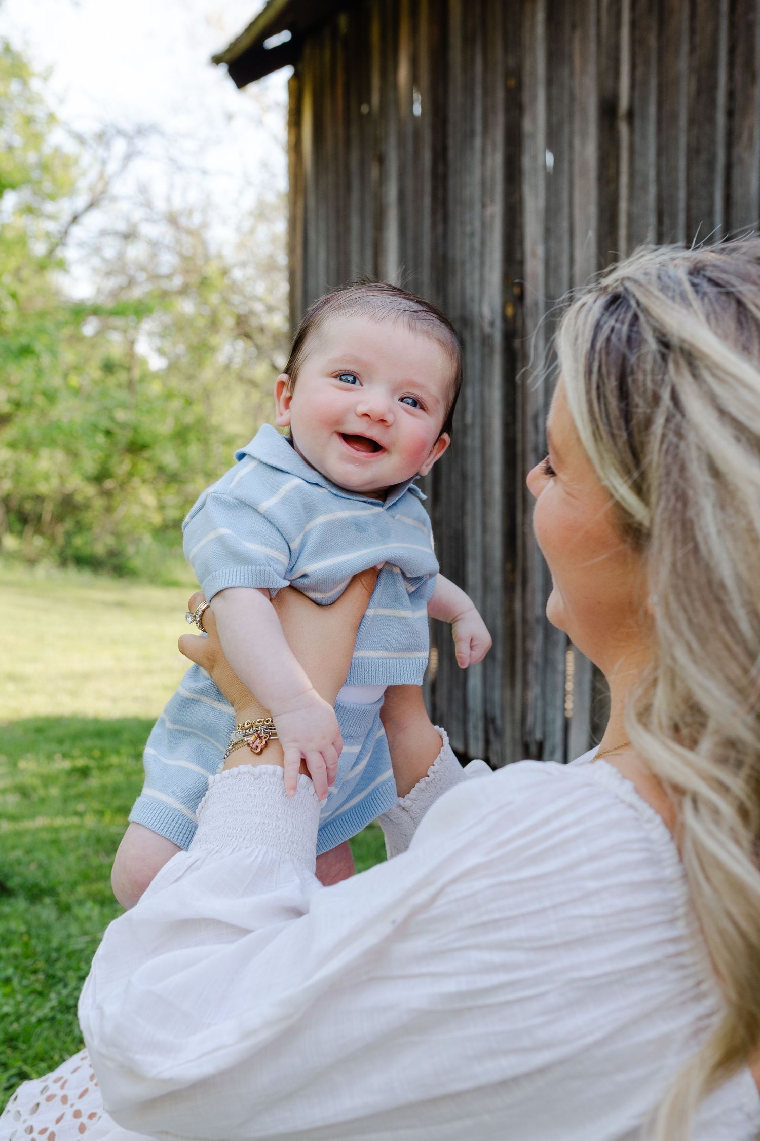 Laughing baby in blue striped outfit being held up by blonde mother near a rustic barn.