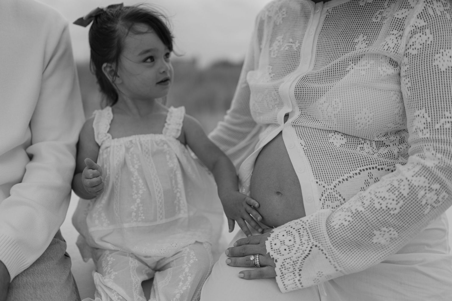 Black and white photo of a young girl in white dress touching pregnant woman's belly during a maternity photoshoot.