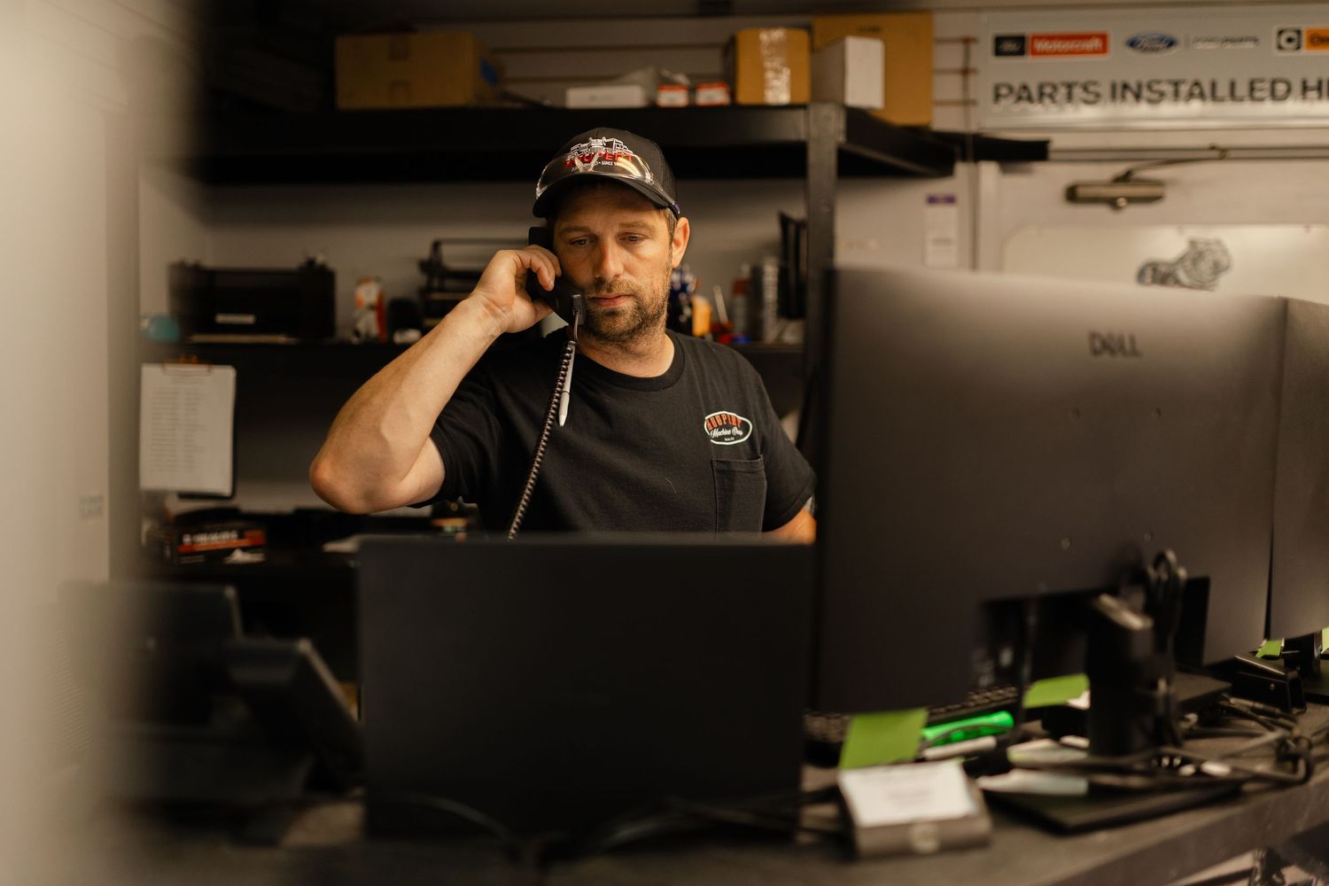 Auto mechanic talking on phone at a computer workstation in a busy garage repair shop.