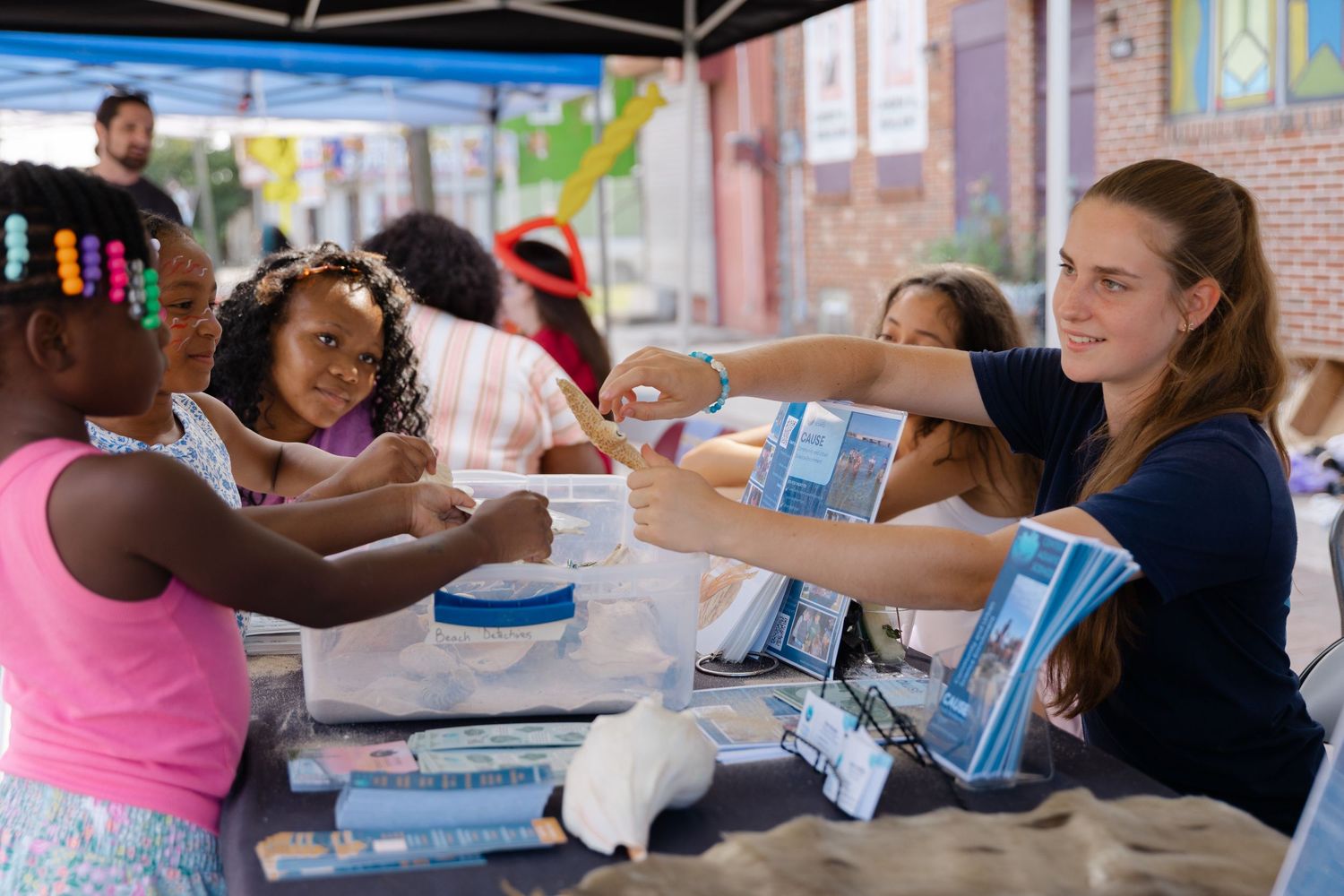 A volunteer woman hands crafts supplies to children at an outdoor community event under a tent on a sunny day.