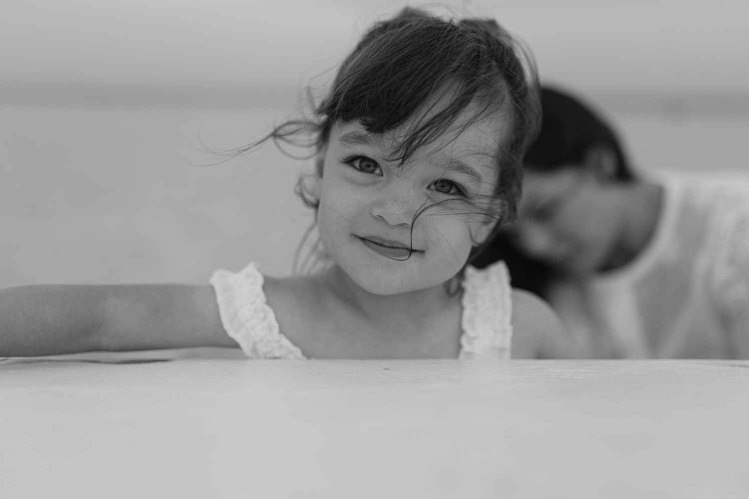 A smiling young girl with dark hair leans on a surface in a black and white photo, with a blurred figure behind her.