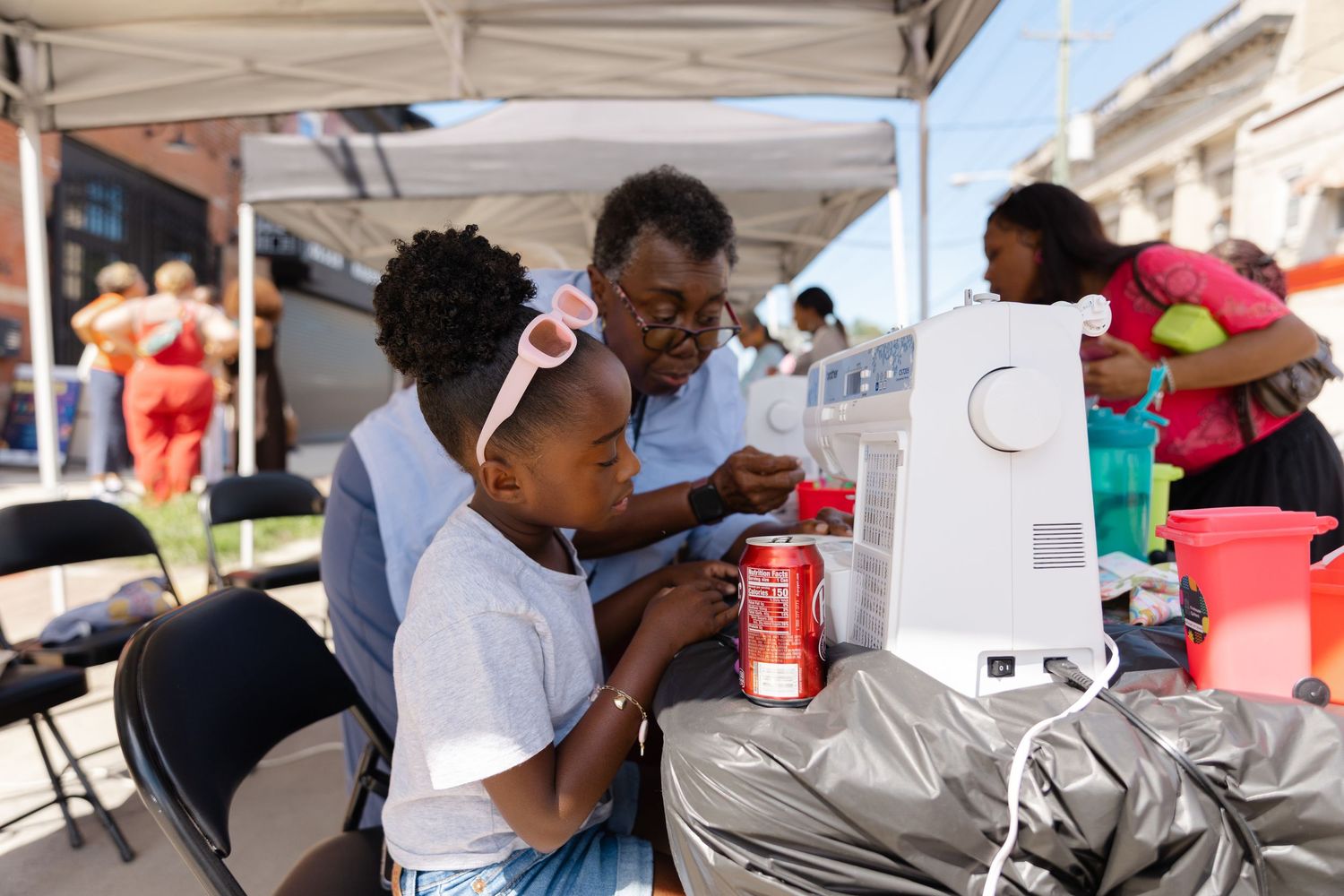 An elderly woman and young girl share a moment at an outdoor community event, with a Coca-Cola can on the table.