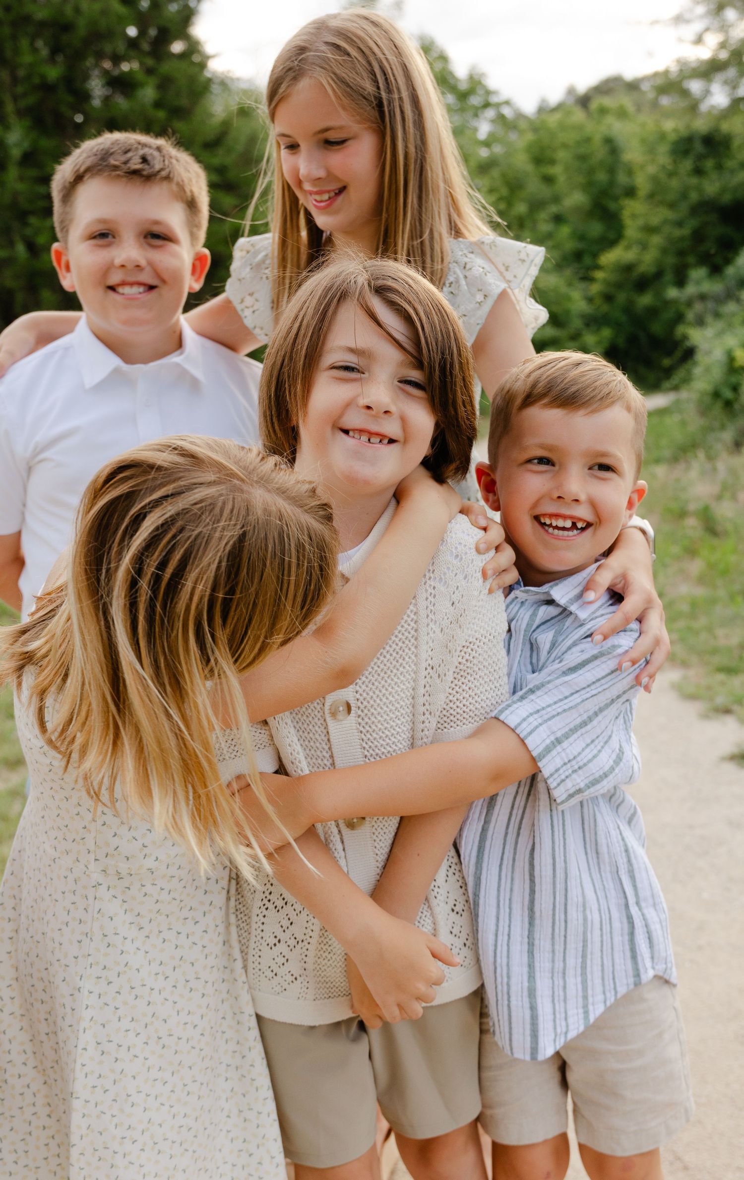 Five children hugging and laughing together on a sunny outdoor dirt path.