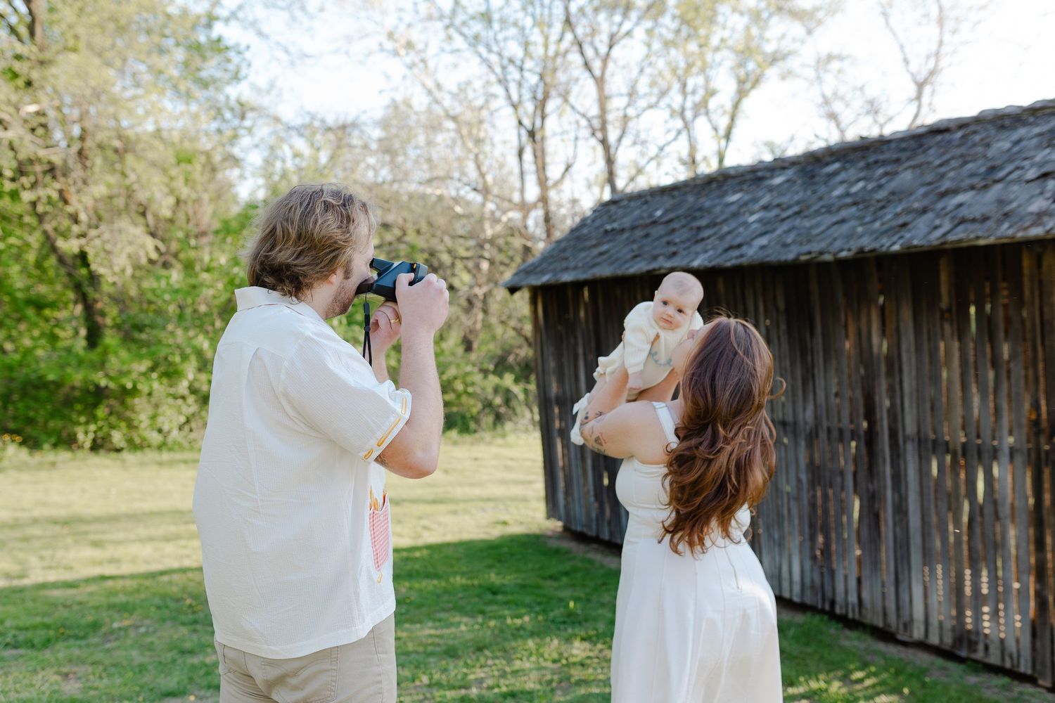 Photographer capturing mother holding baby near rustic wooden barn outdoors.
