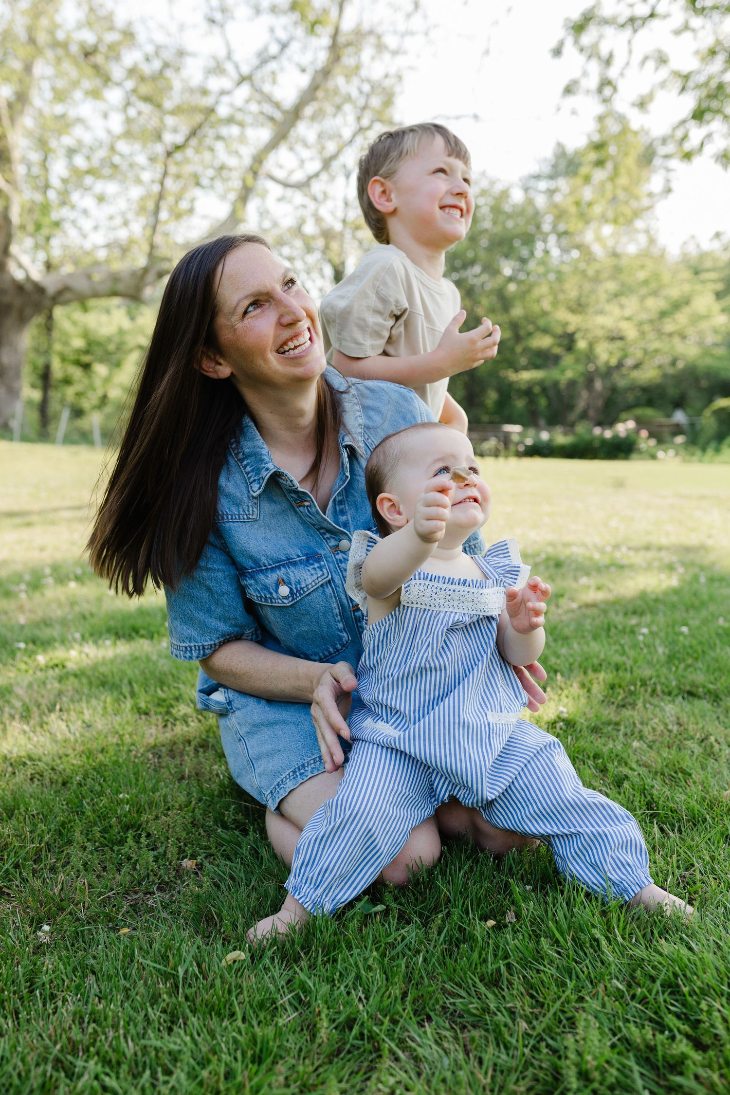 Smiling mother sitting on grass with two young children in a sunny green park.