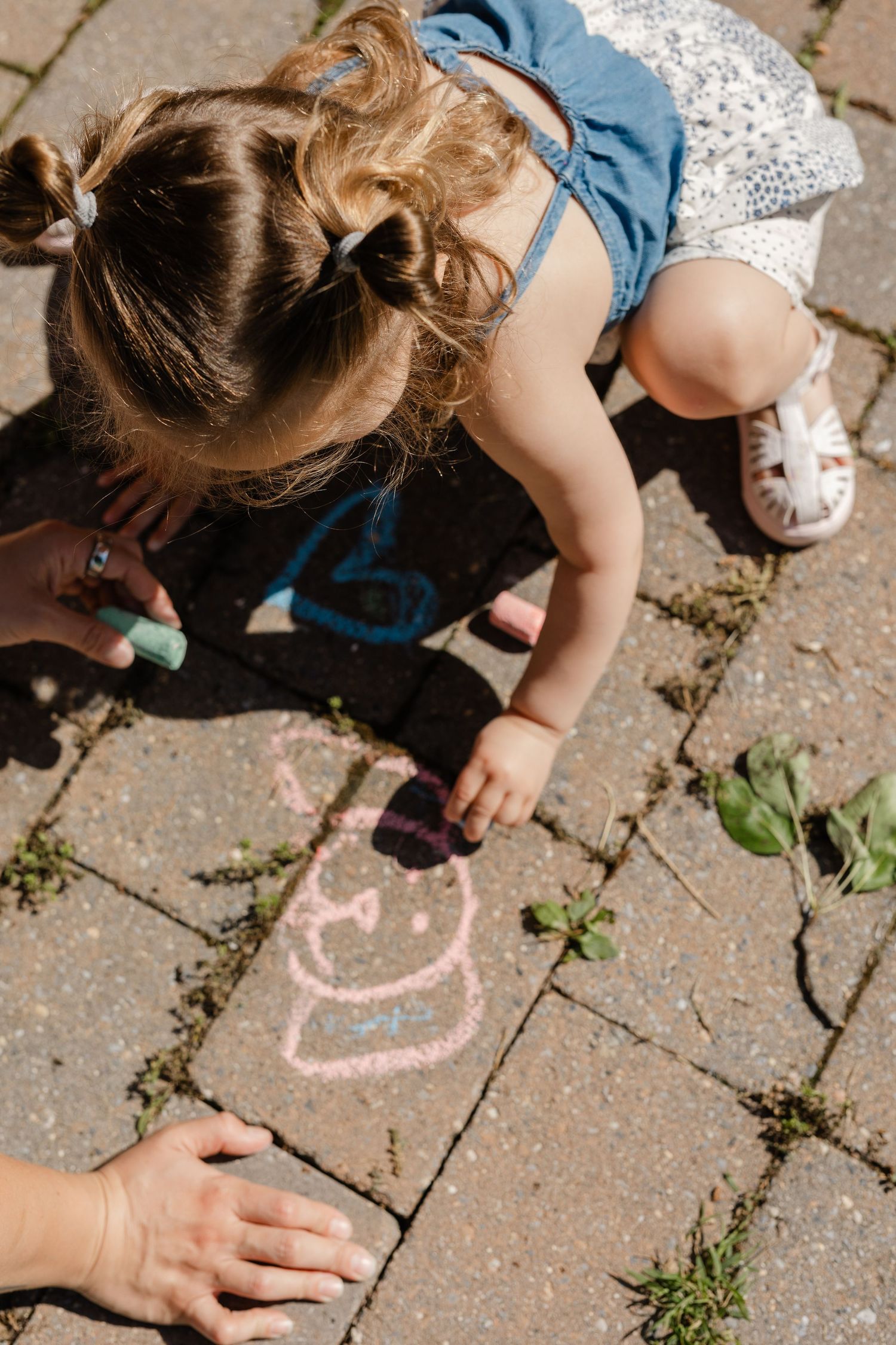 Young toddler girl drawing with colorful chalk on a stone patio outdoors in summer sunshine.