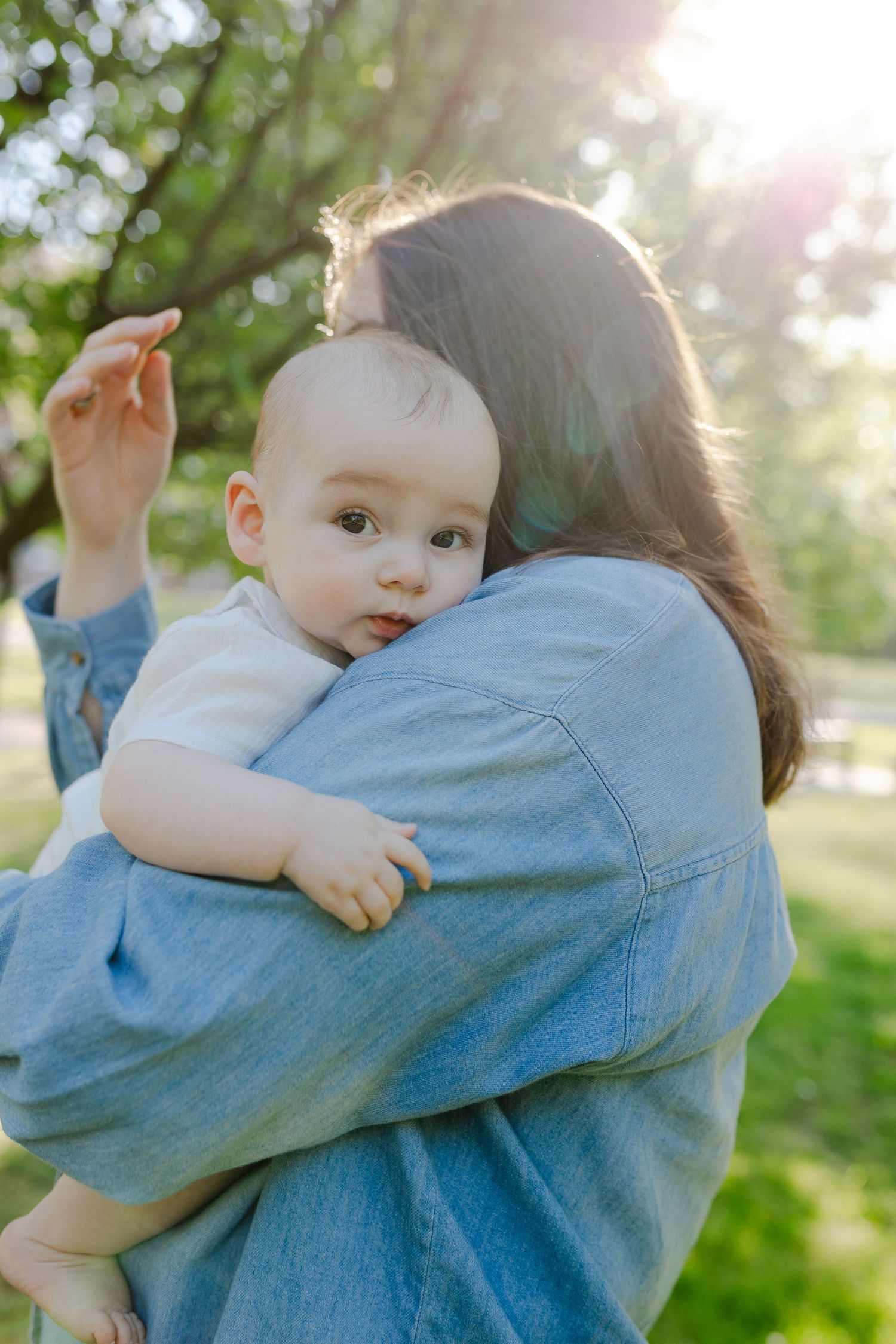 Mother holding baby against her shoulder in sunny park, baby looking curiously at camera.
