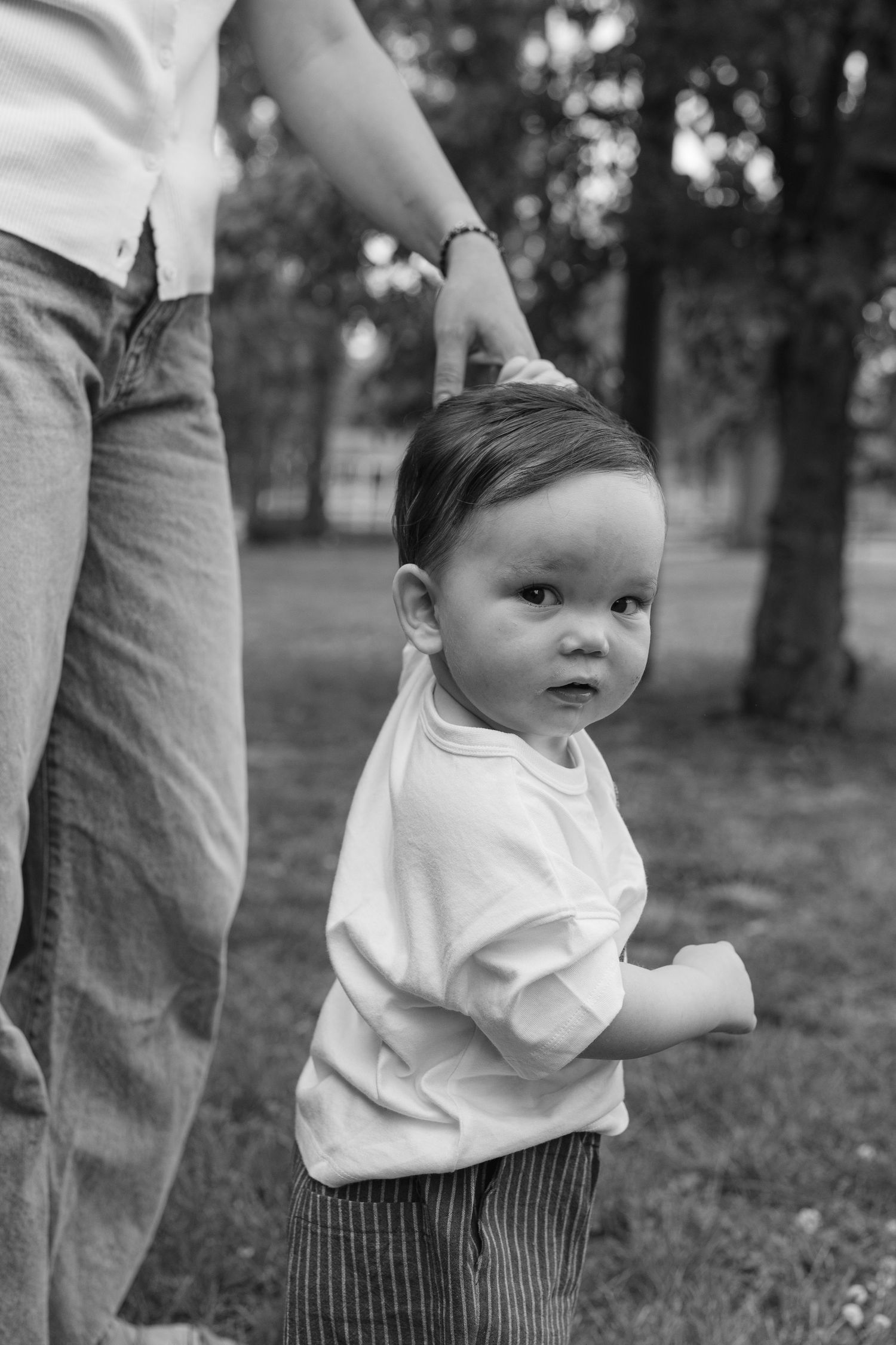 Black and white photo of toddler learning to walk while holding parent's hand outdoors.