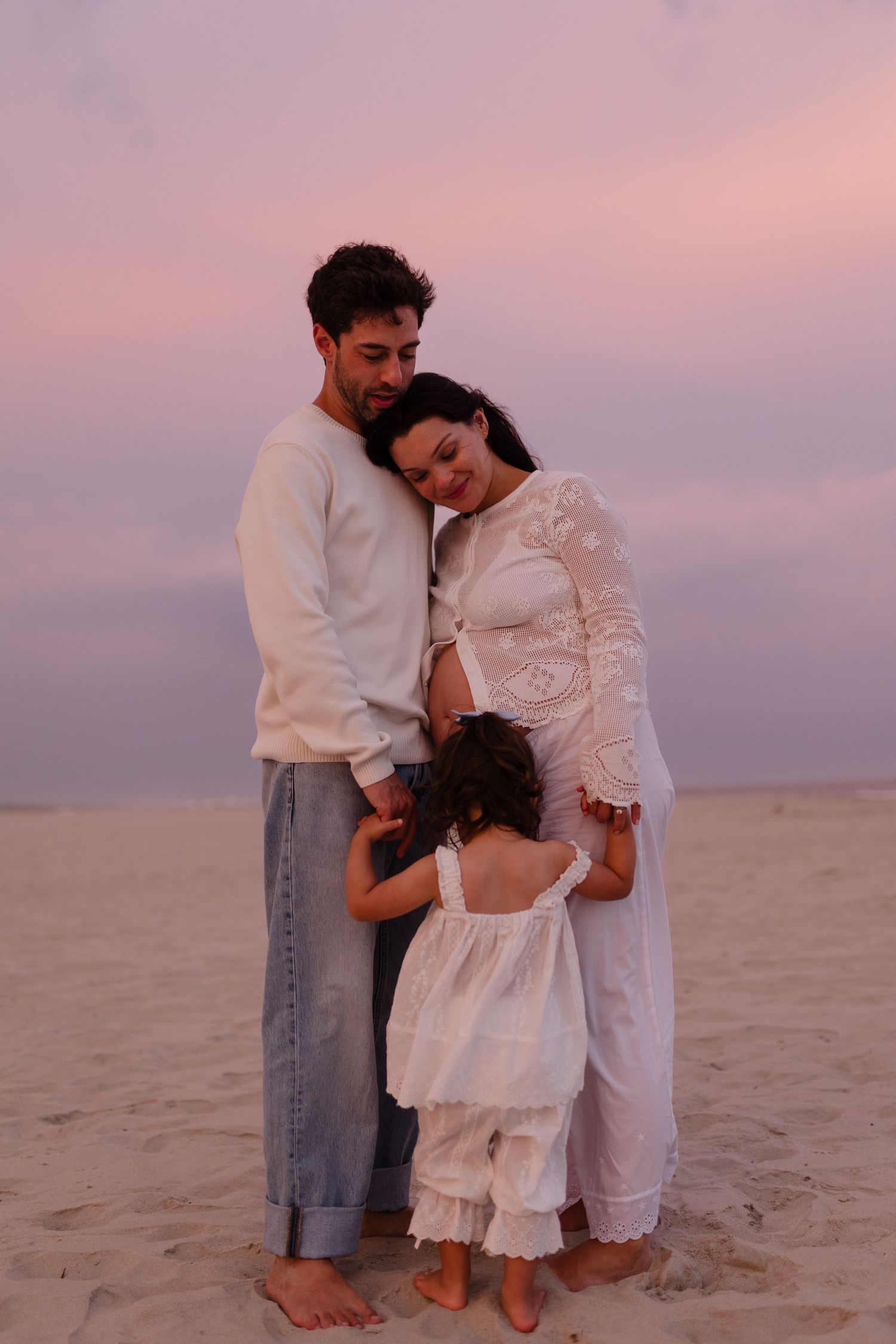 A family of three shares a tender embrace on a sandy beach under a pink sunset sky.