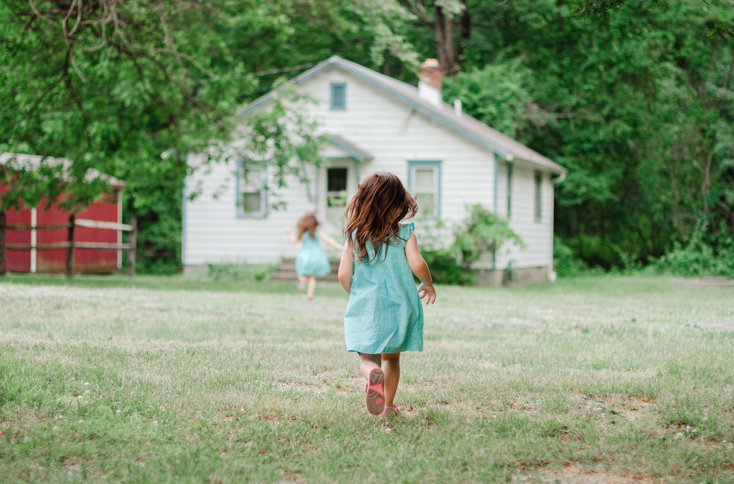Two young girls in matching teal dresses running toward a white farmhouse surrounded by lush green trees.