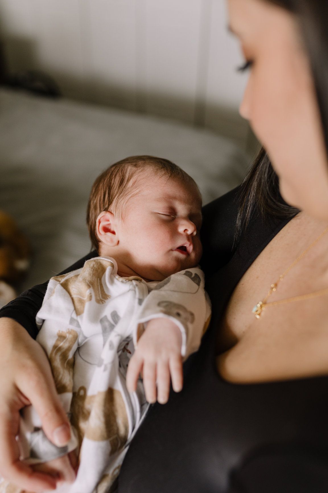A dark-haired mother gazes lovingly at her sleeping newborn baby wrapped in a patterned blanket.