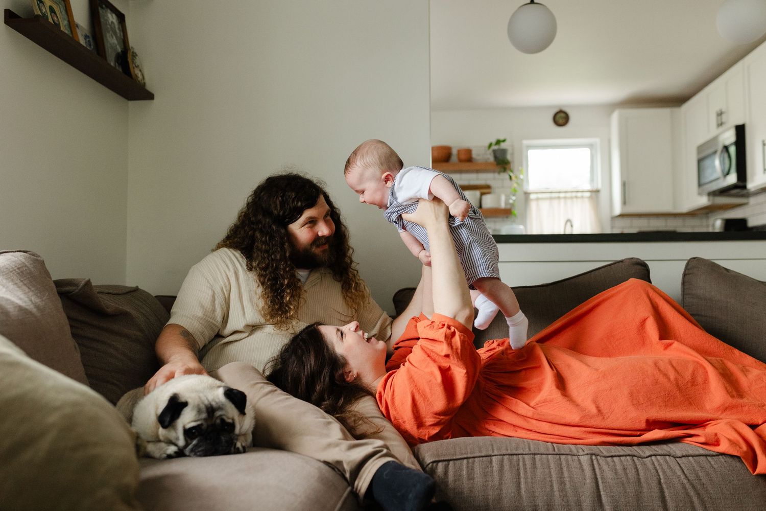 Young family with curly-haired father, mother in orange dress, baby, and pug relaxing on a couch.