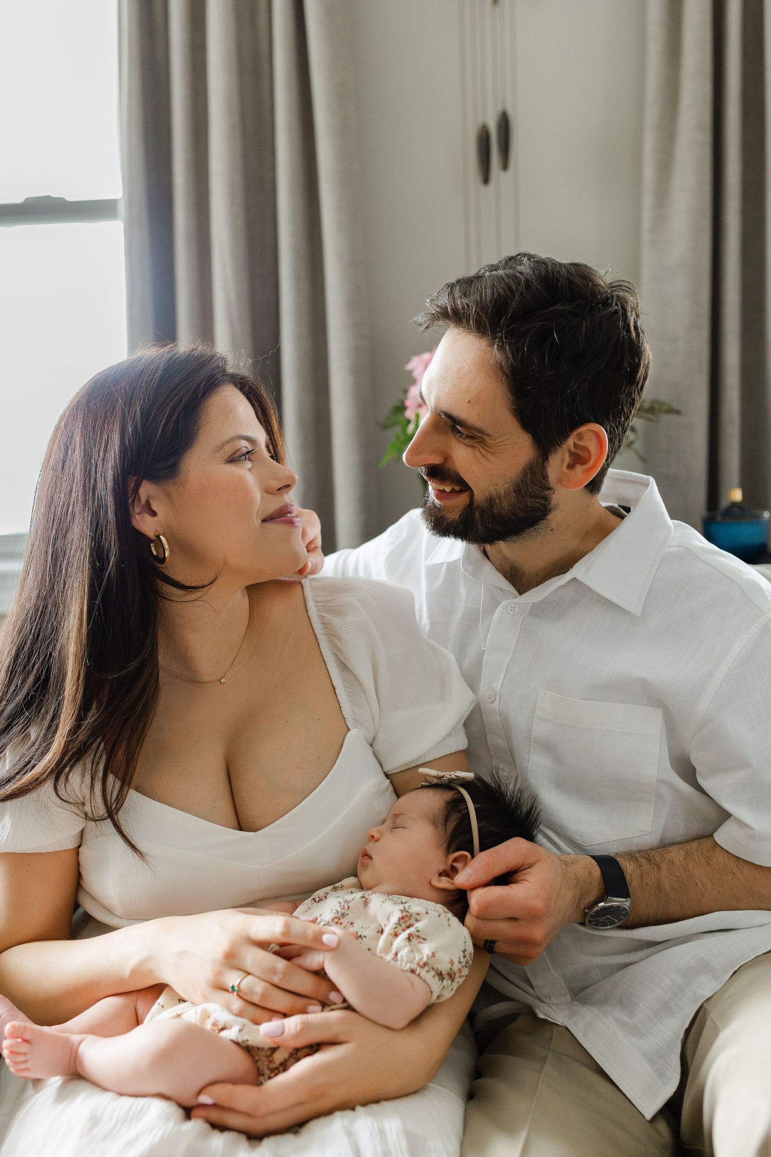 Happy couple in white outfits sitting together smiling while mother holds sleeping newborn baby.