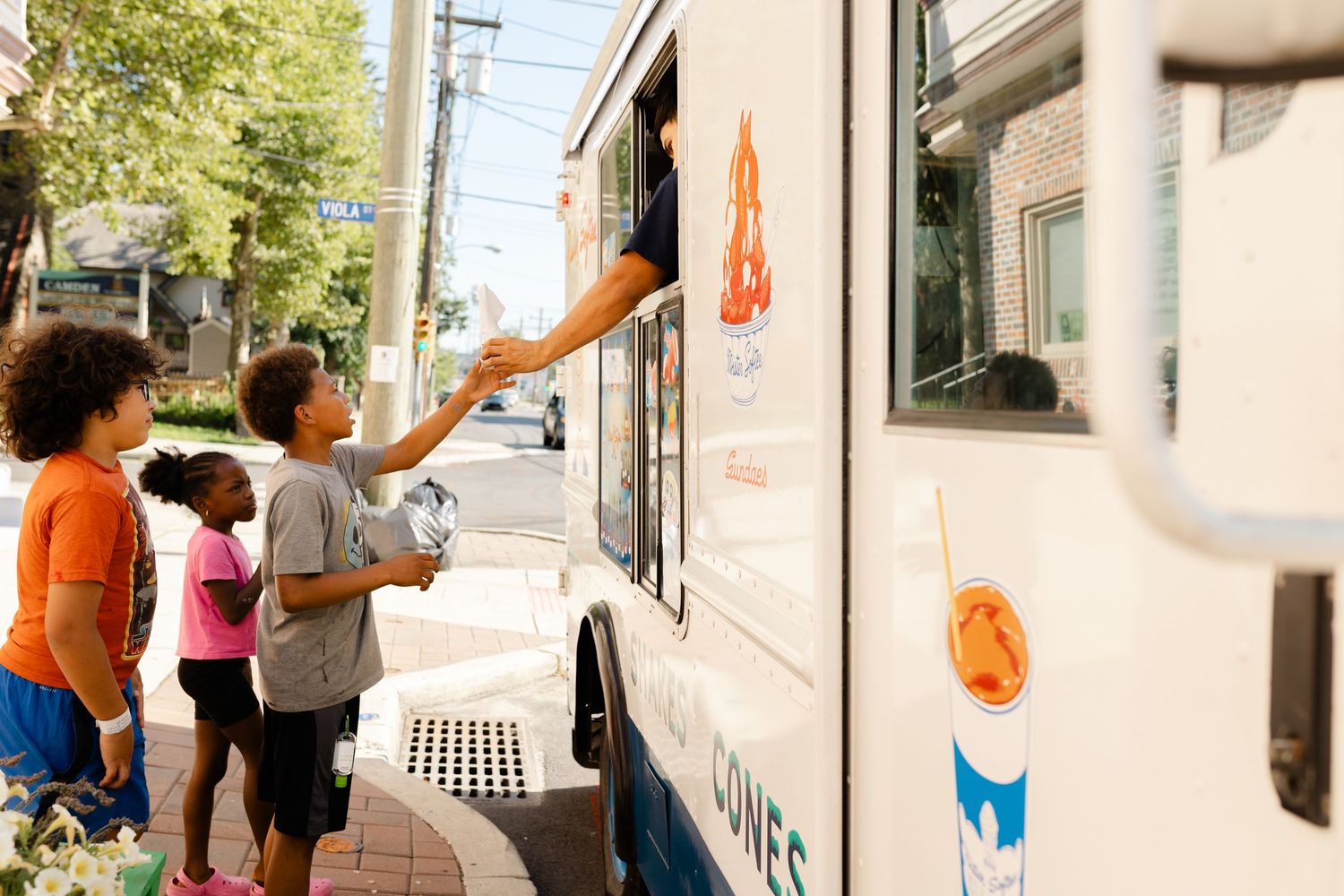 Children buying ice cream from a street vendor truck on a sunny urban sidewalk.