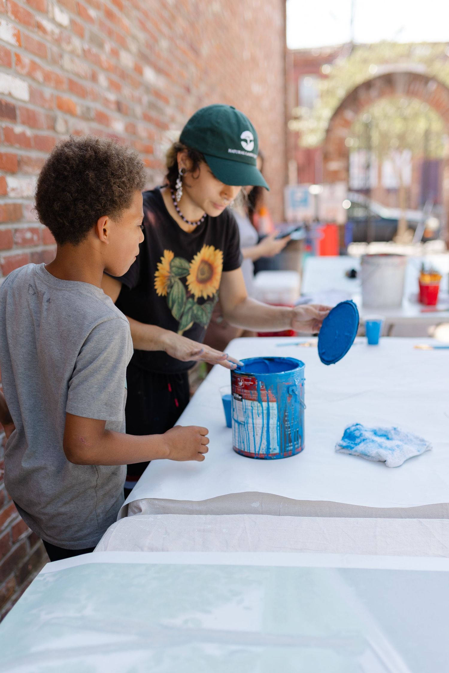 Boy watches woman in sunflower shirt open blue paint can at outdoor art table against brick wall.