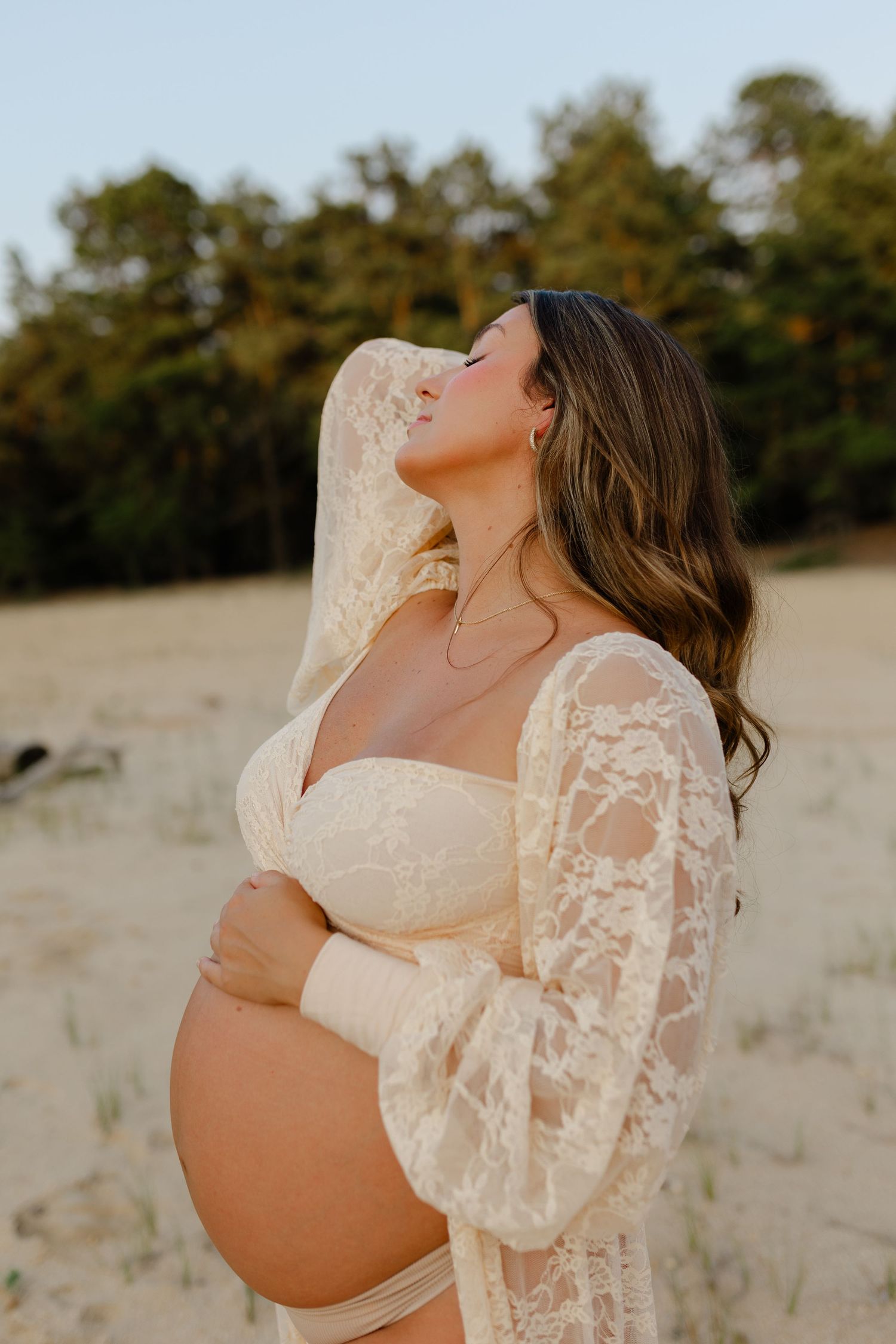 Pregnant woman in cream lace robe tilts head back holding baby bump on sandy beach at dusk.