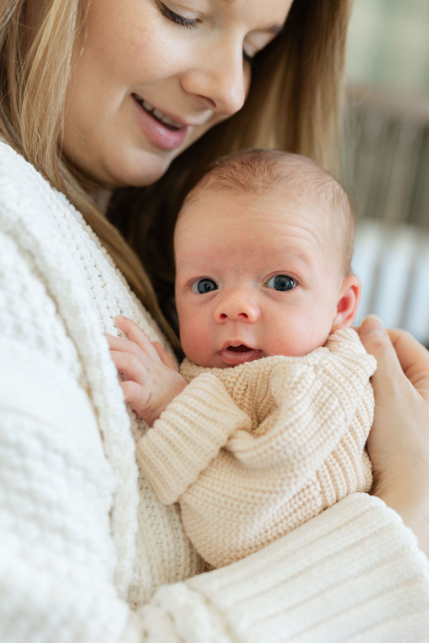 A smiling blonde mother in a white knit sweater cradles her wide-eyed newborn baby.