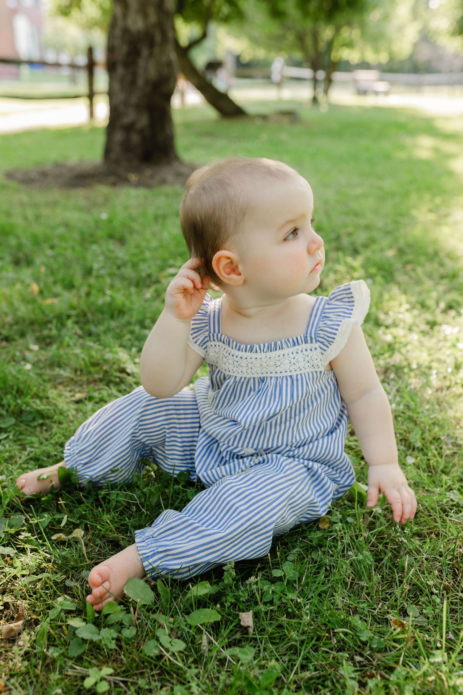 Adorable baby in blue striped romper sitting on green grass under a tree outdoors.