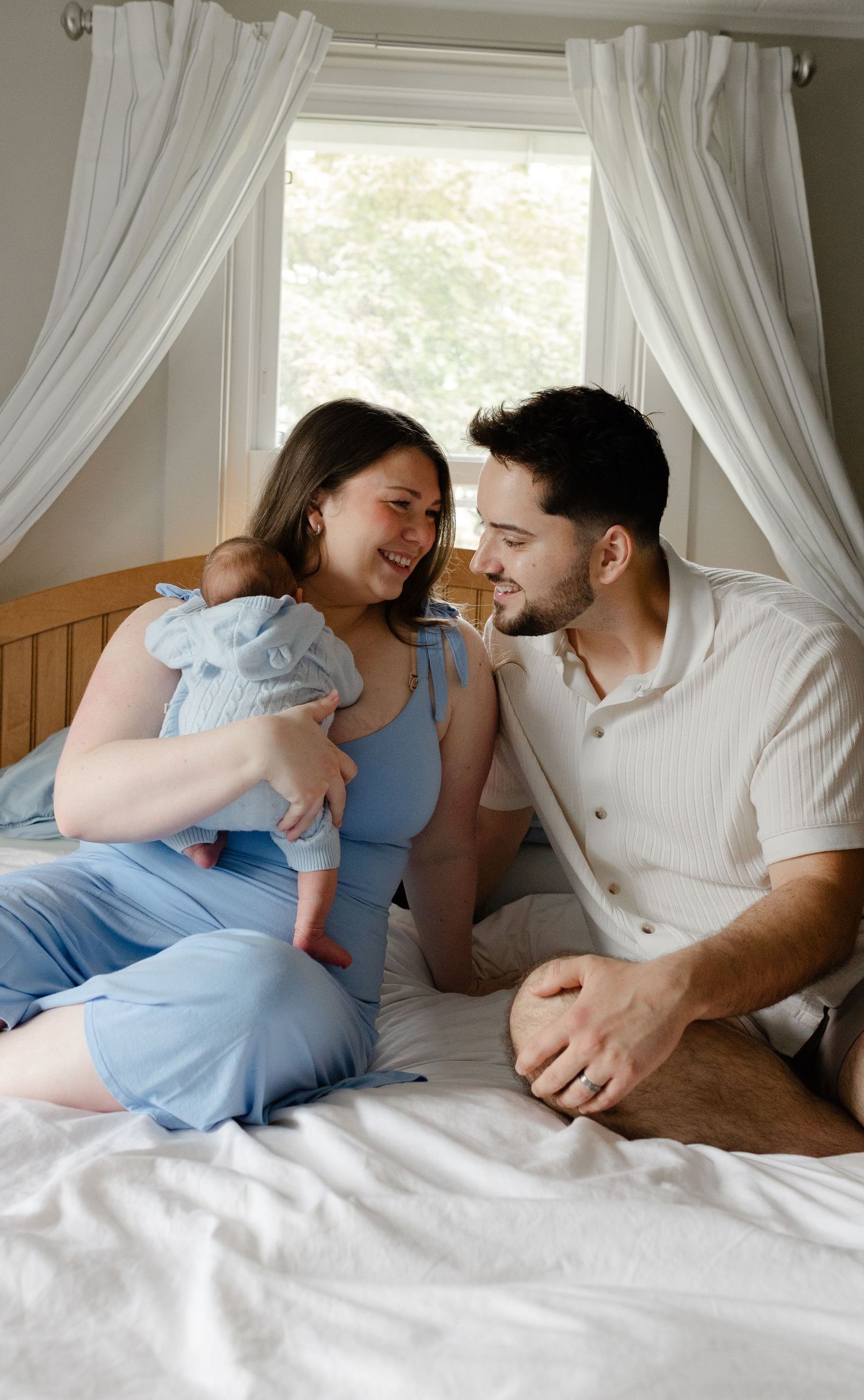 Happy couple sitting on bed smiling at each other while mother holds newborn baby in blue outfit.