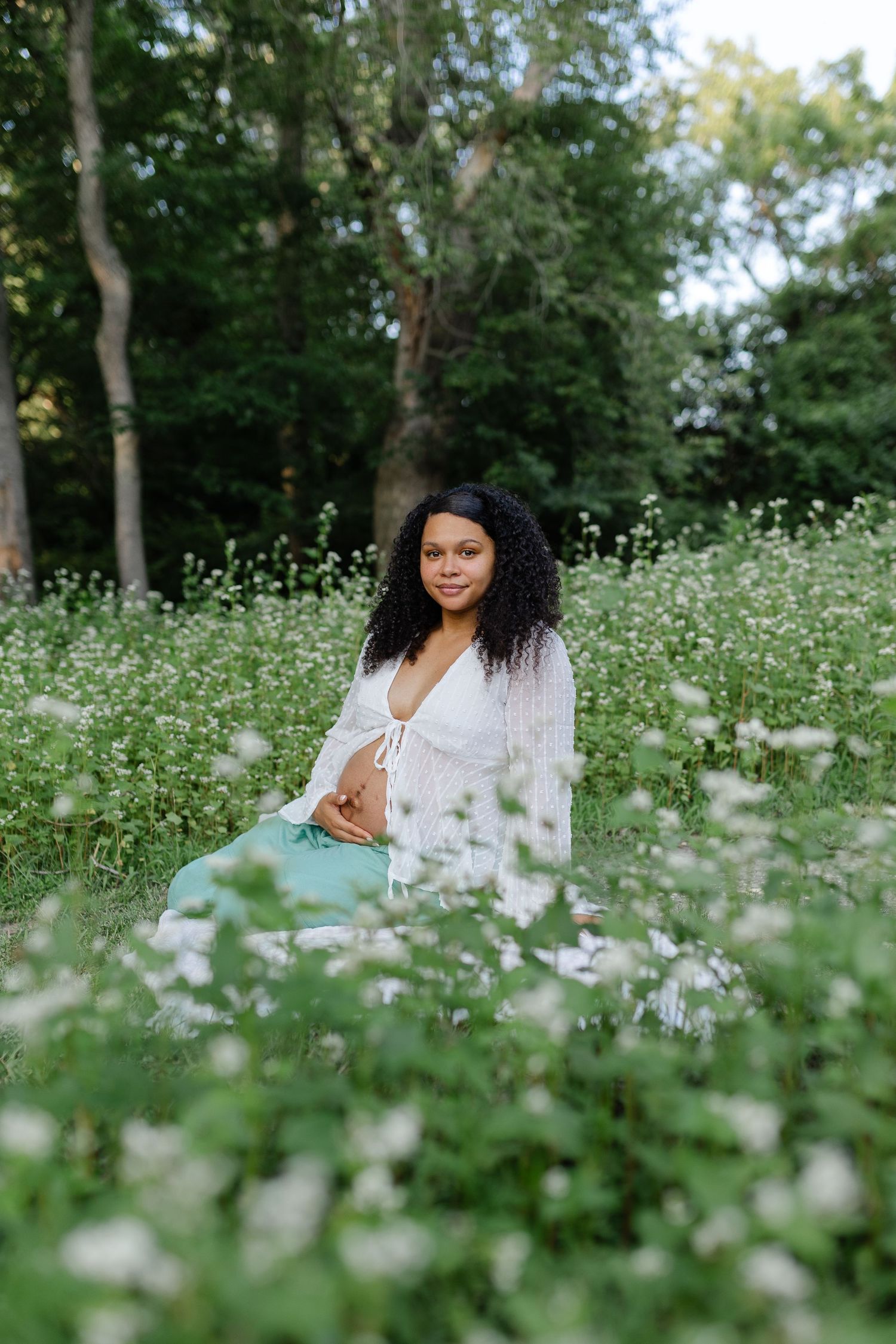 Smiling pregnant woman in white top and teal skirt sitting in field of white flowers.