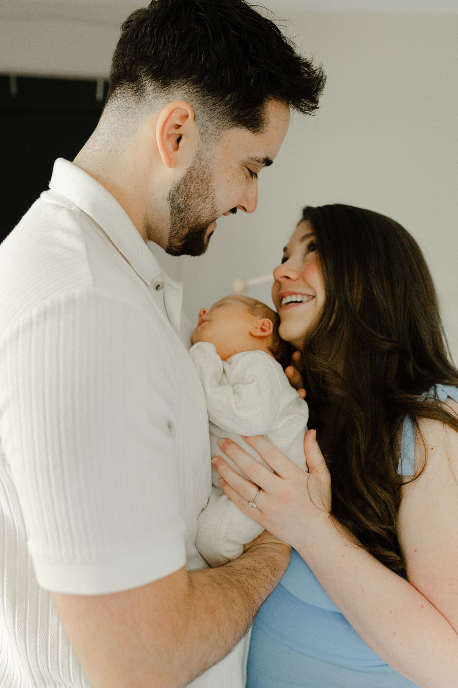 New parents smiling and cradling their sleeping newborn baby, father in white shirt, mother in blue dress.